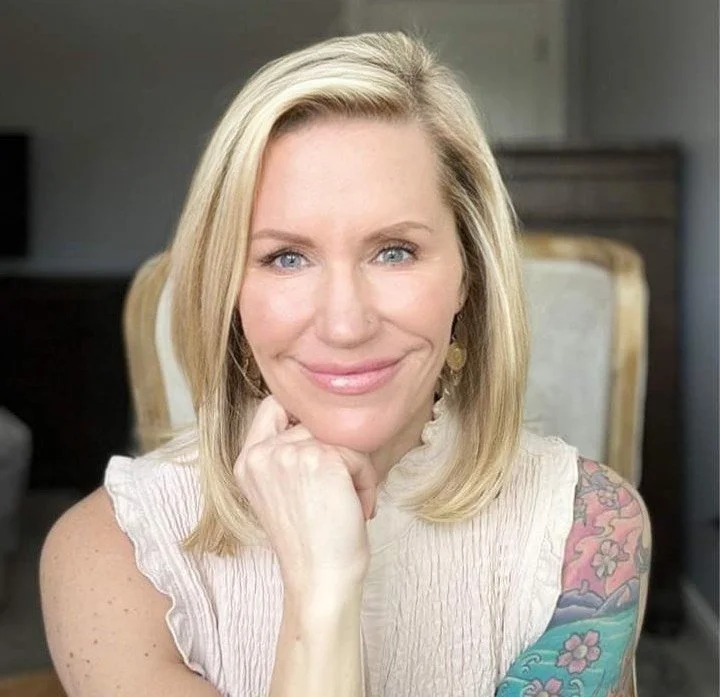 Close-up of a smiling blonde woman with blue eyes, wearing a white sleeveless top, resting her chin on her hand, with a colorful tattoo on her arm, in a home setting.