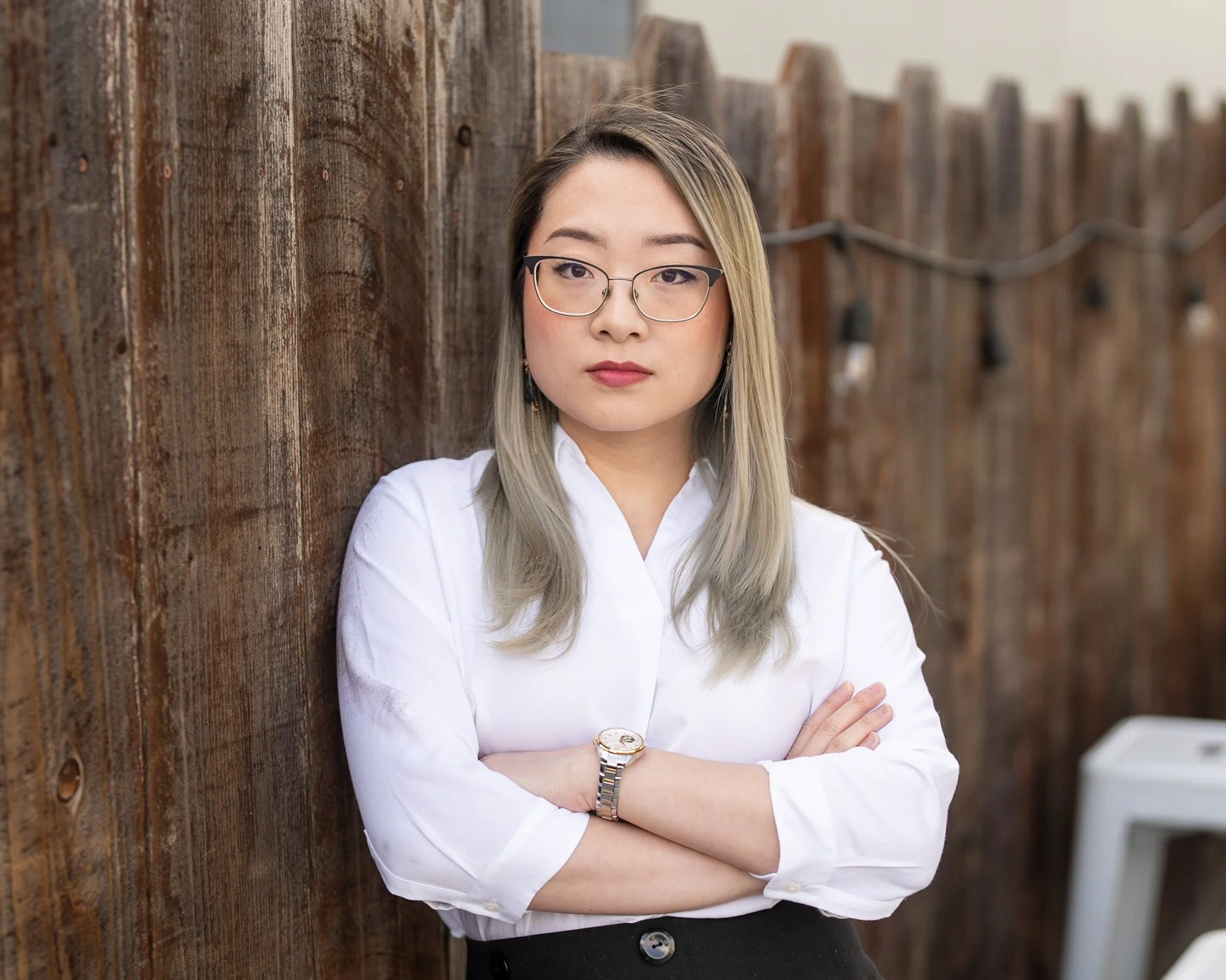 A woman with glasses and shoulder-length blonde hair, wearing a white blouse and black pants, standing with arms crossed and leaning against a wooden fence outdoors.