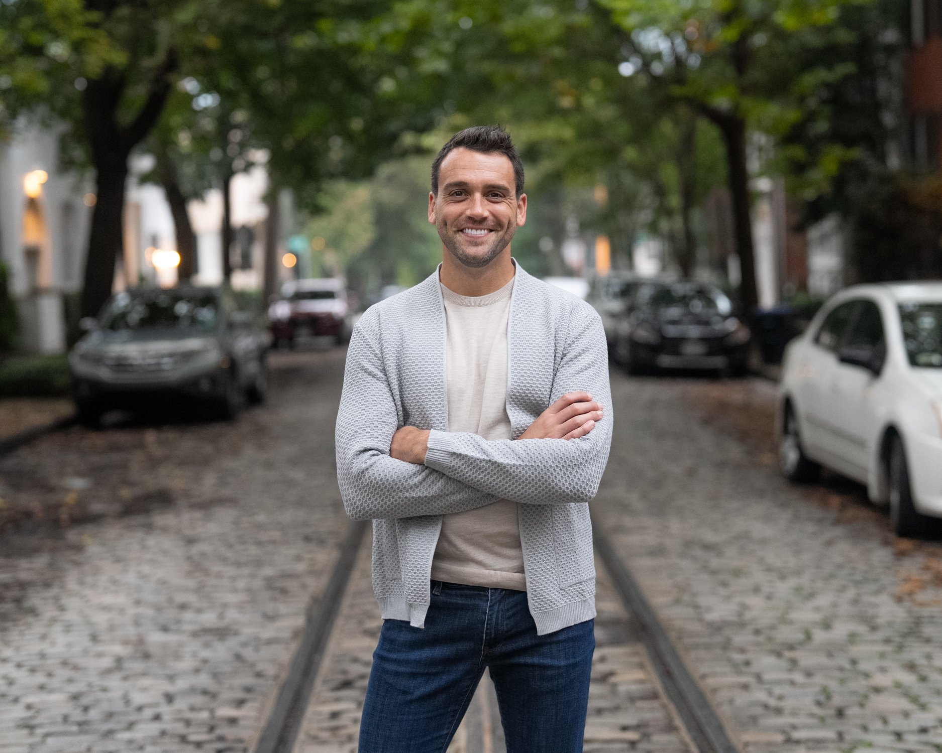 A smiling man standing with crossed arms on a cobblestone street with parked cars and trees in the background.