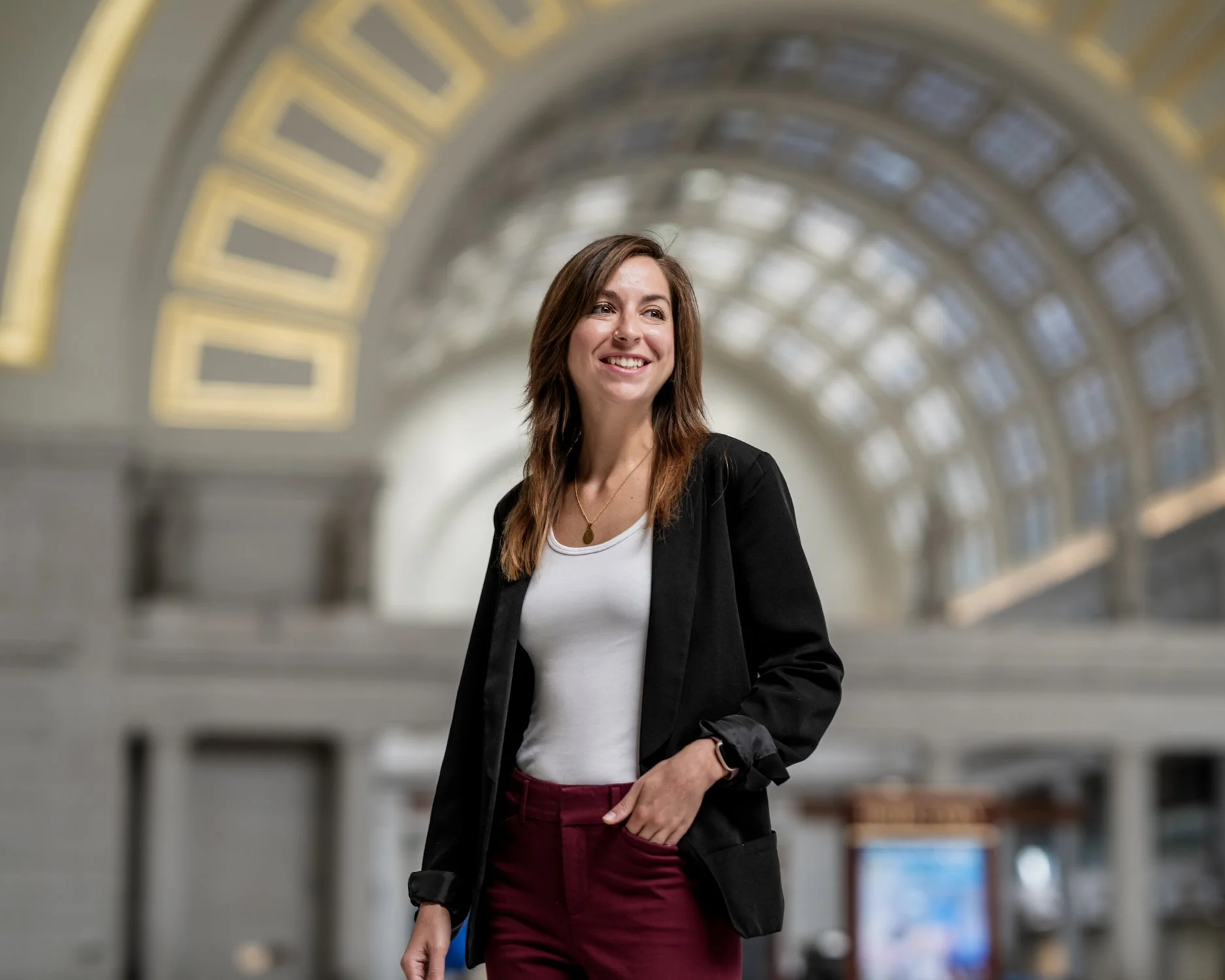 A smiling woman wearing a black blazer and maroon pants walking in a large, historic train station with arched windows and decorative yellow accents.