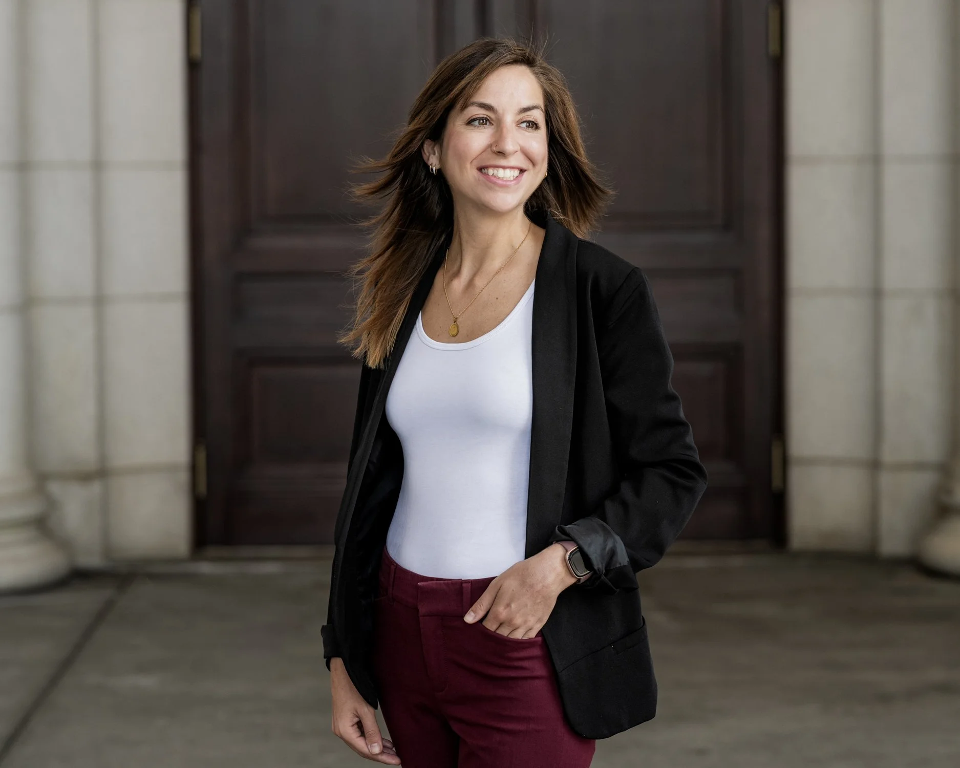 A woman smiling and posing outdoors in front of a large wooden door, wearing a black blazer, white t-shirt, burgundy pants, and a gold necklace.