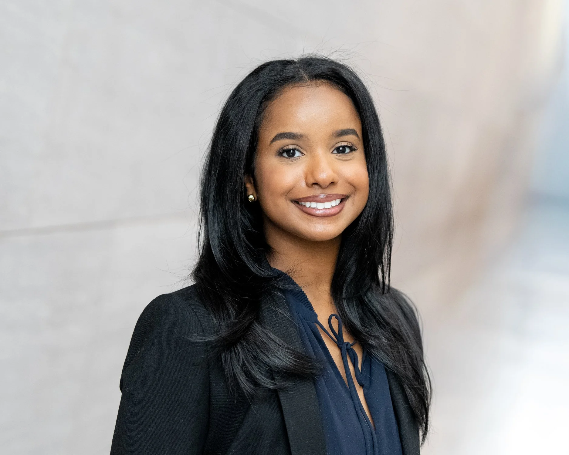 A professional woman with long black hair, smiling, wearing a dark blazer and a navy blouse, standing in front of a plain light-colored wall.