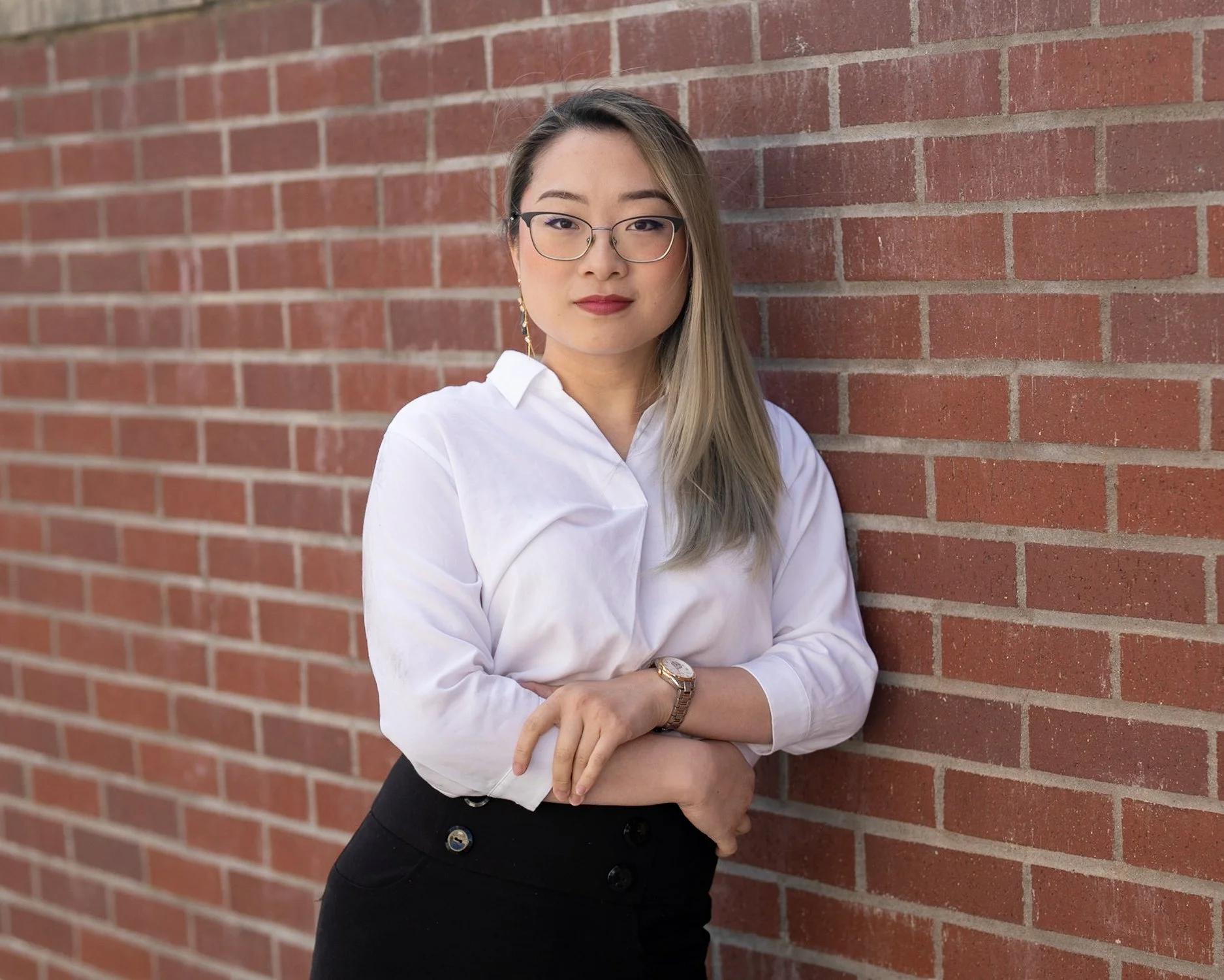 A woman with glasses and long blonde hair standing against a red brick wall, wearing a white blouse and black skirt.