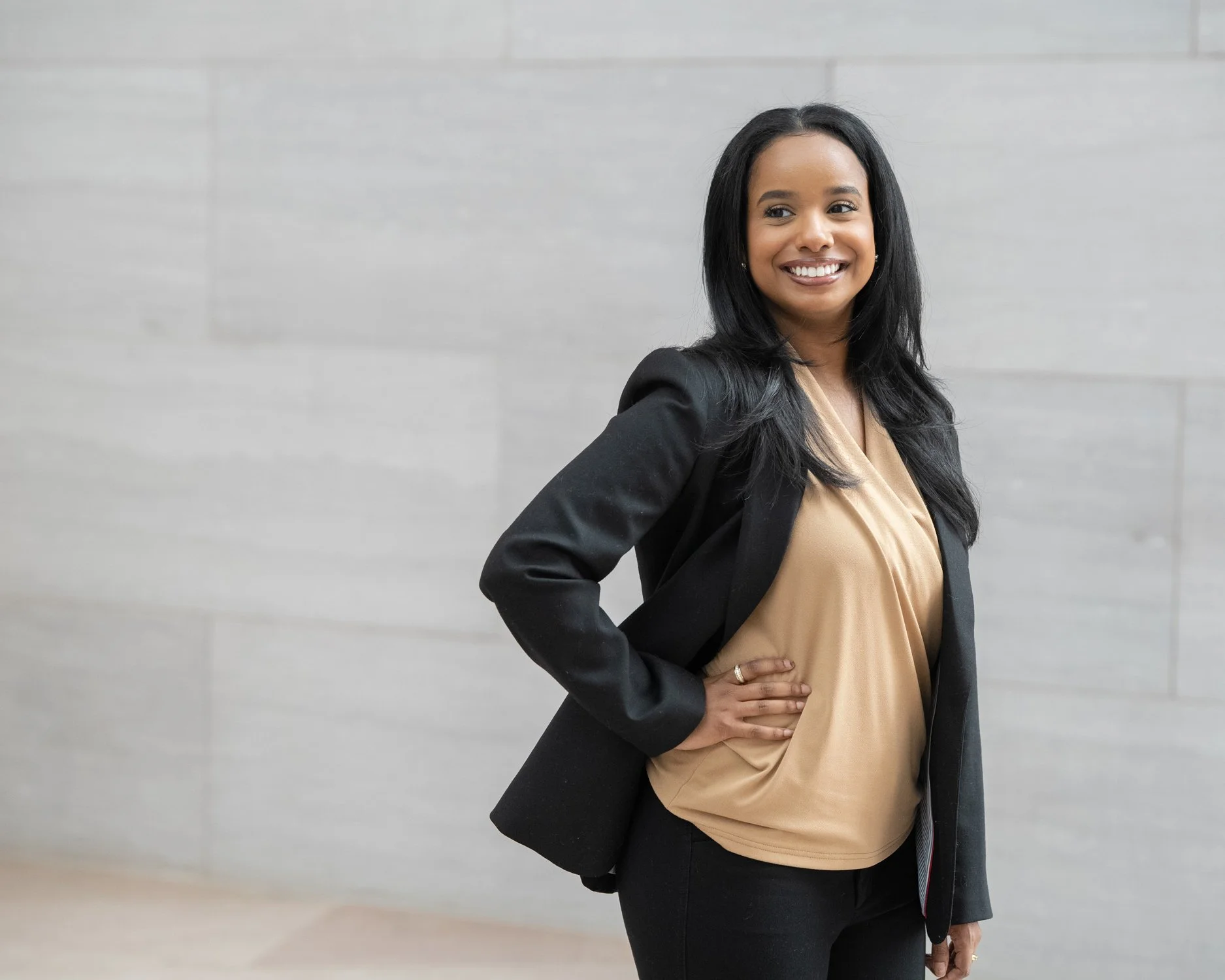 A smiling woman wearing a beige blouse and black blazer, standing against a light gray wall.