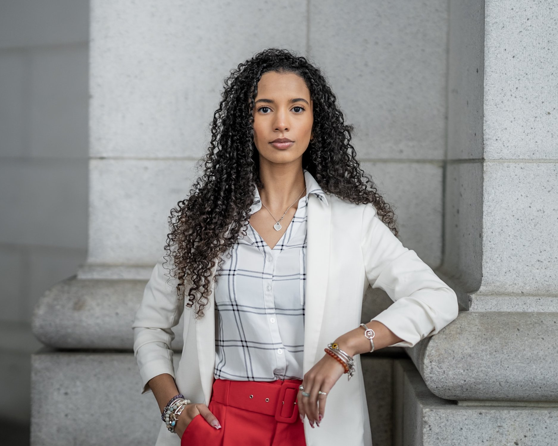 A woman with long curly black hair standing against a stone building wall, wearing a white blazer, plaid shirt, and red pants, accessorized with jewelry.