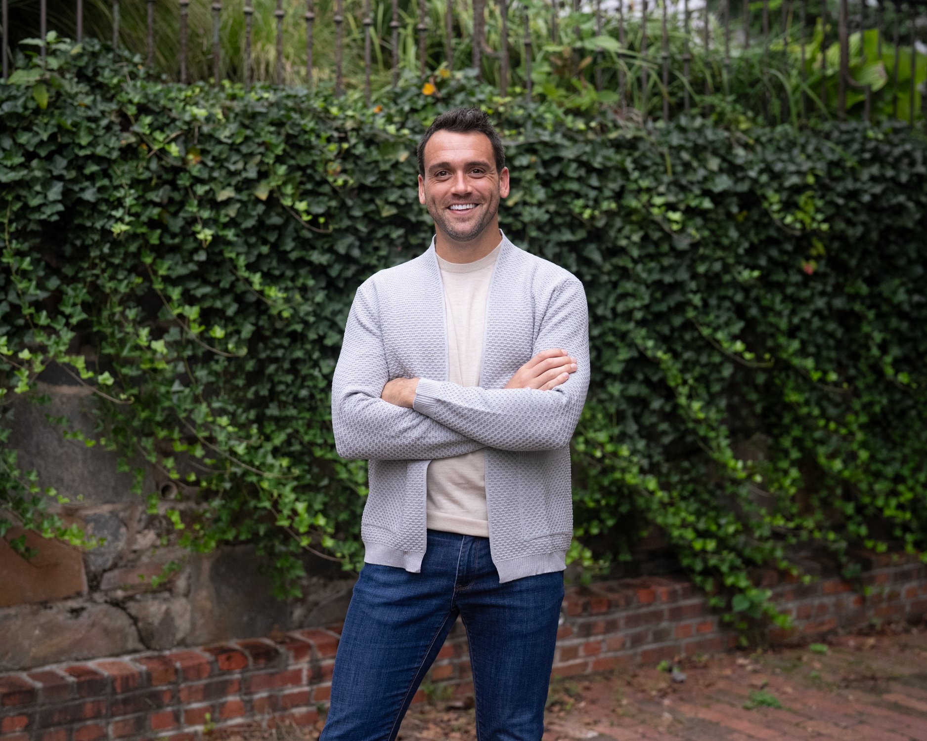 A man stands outdoors in front of green ivy and brick wall, smiling with arms crossed, wearing a white t-shirt, gray cardigan, and blue jeans.
