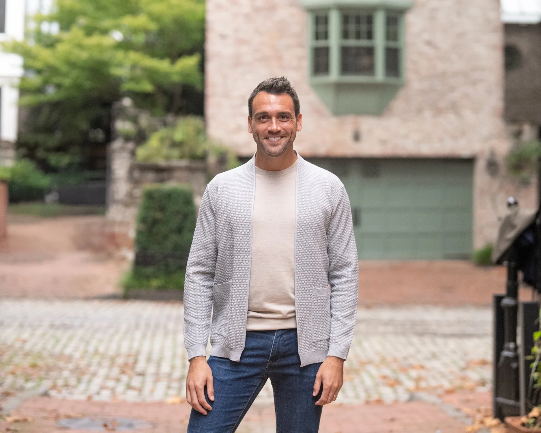 A smiling man standing outdoors on a brick and stone driveway with a house and greenery in the background.
