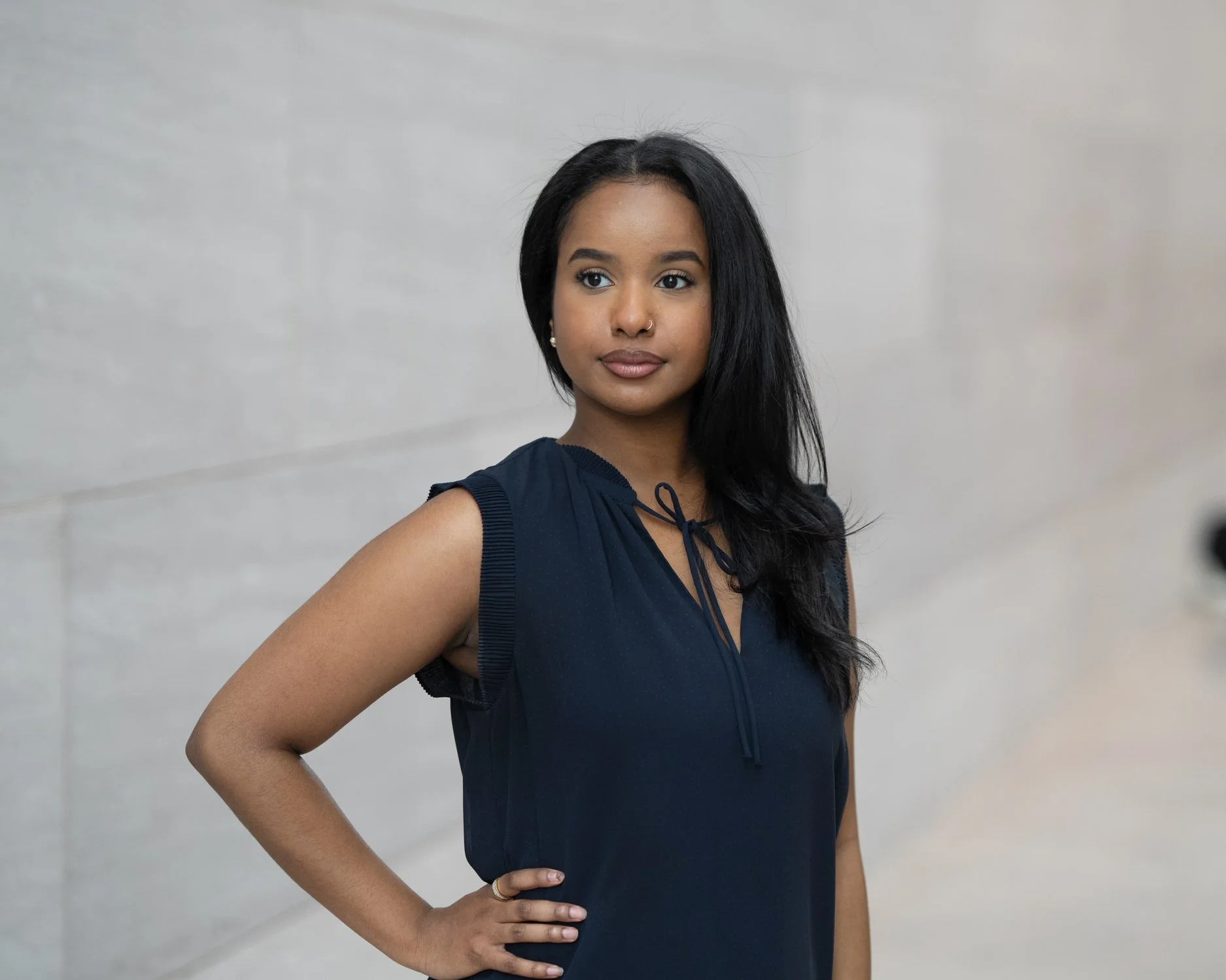A woman with long black hair wearing a sleeveless navy blue dress, standing against a gray wall, looking to her right with her hand on her hip.