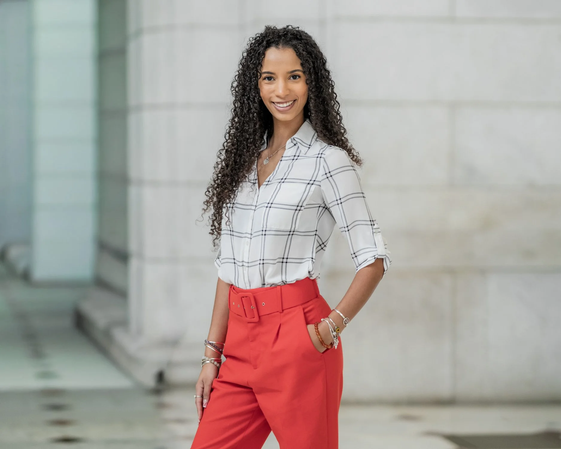 A woman with curly hair smiling, standing outdoors against a modern building wall, wearing a white plaid shirt and red high-waisted pants.