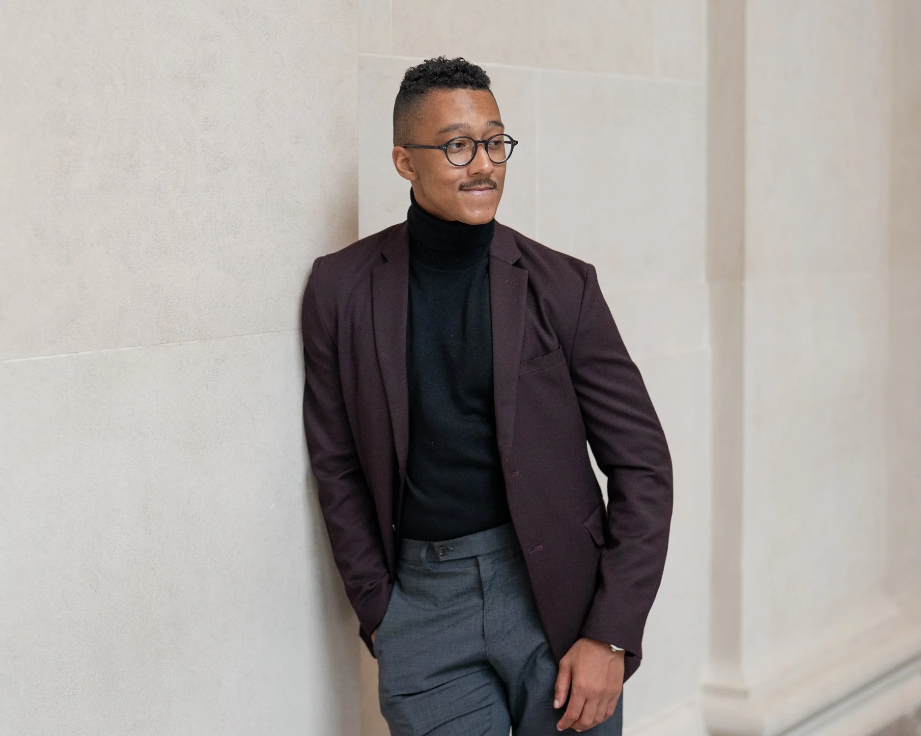 A man in a dark blazer and turtleneck leans against a beige wall, looking to the side with one hand in his pocket.