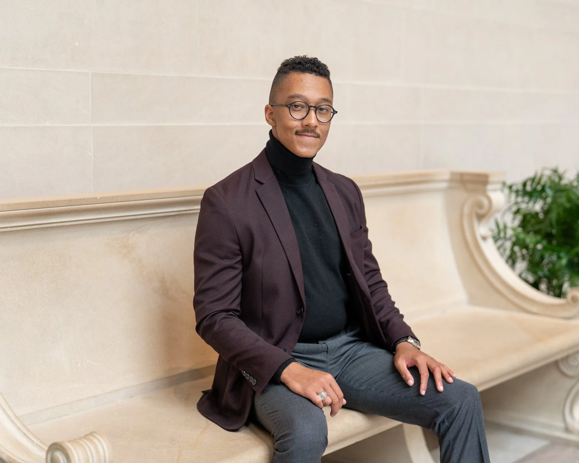 A young man sitting on a beige ornate bench in a formal setting, wearing glasses, a dark turtleneck, a blazer, and gray pants.