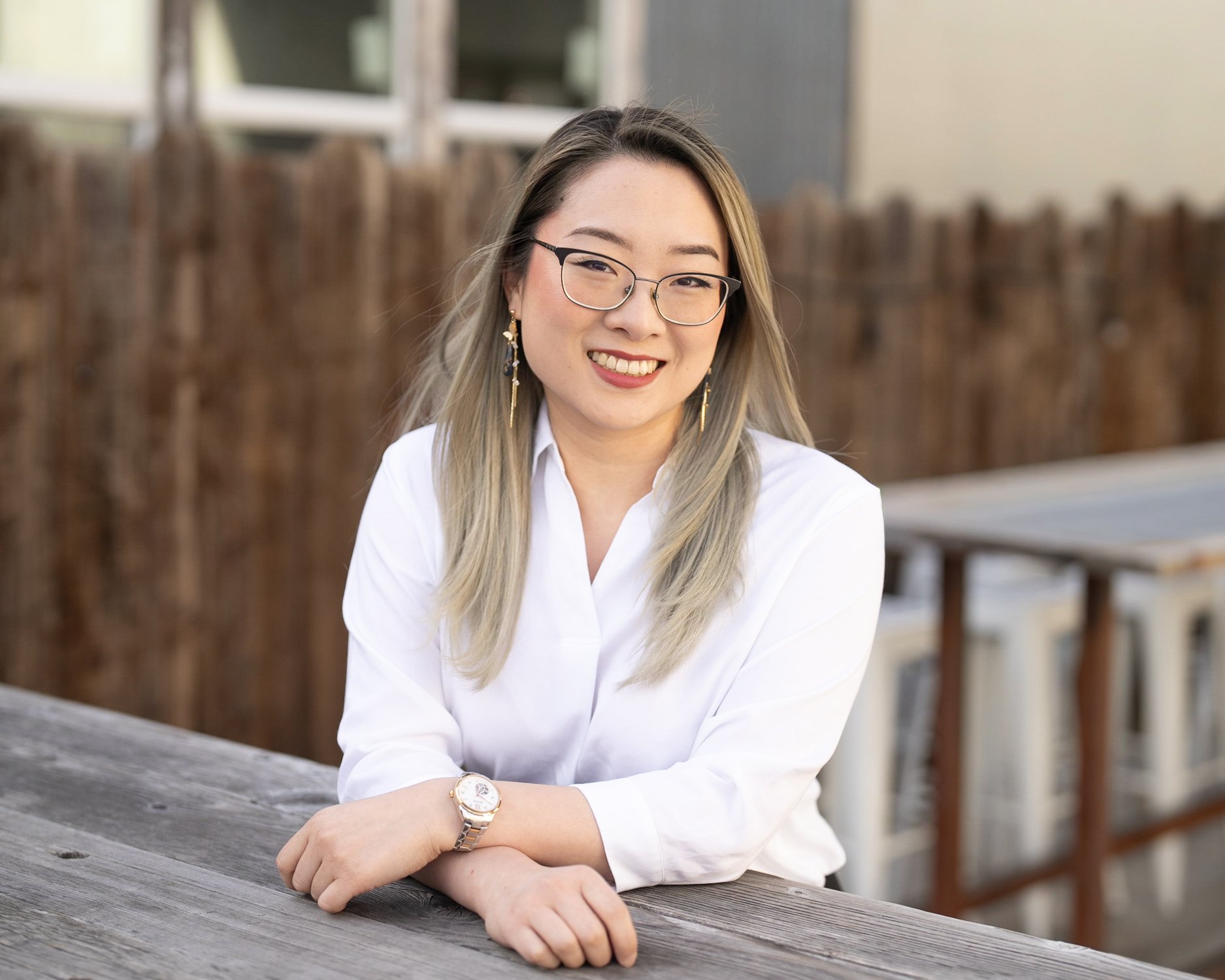 Portrait of a smiling woman with glasses and earrings wearing a white shirt, sitting outdoors at a wooden table.