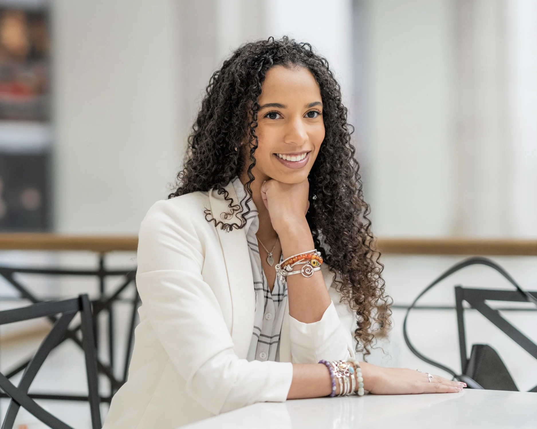 A woman with long curly hair smiles at the camera while sitting at a table in a bright room, wearing a white blazer and jewelry.