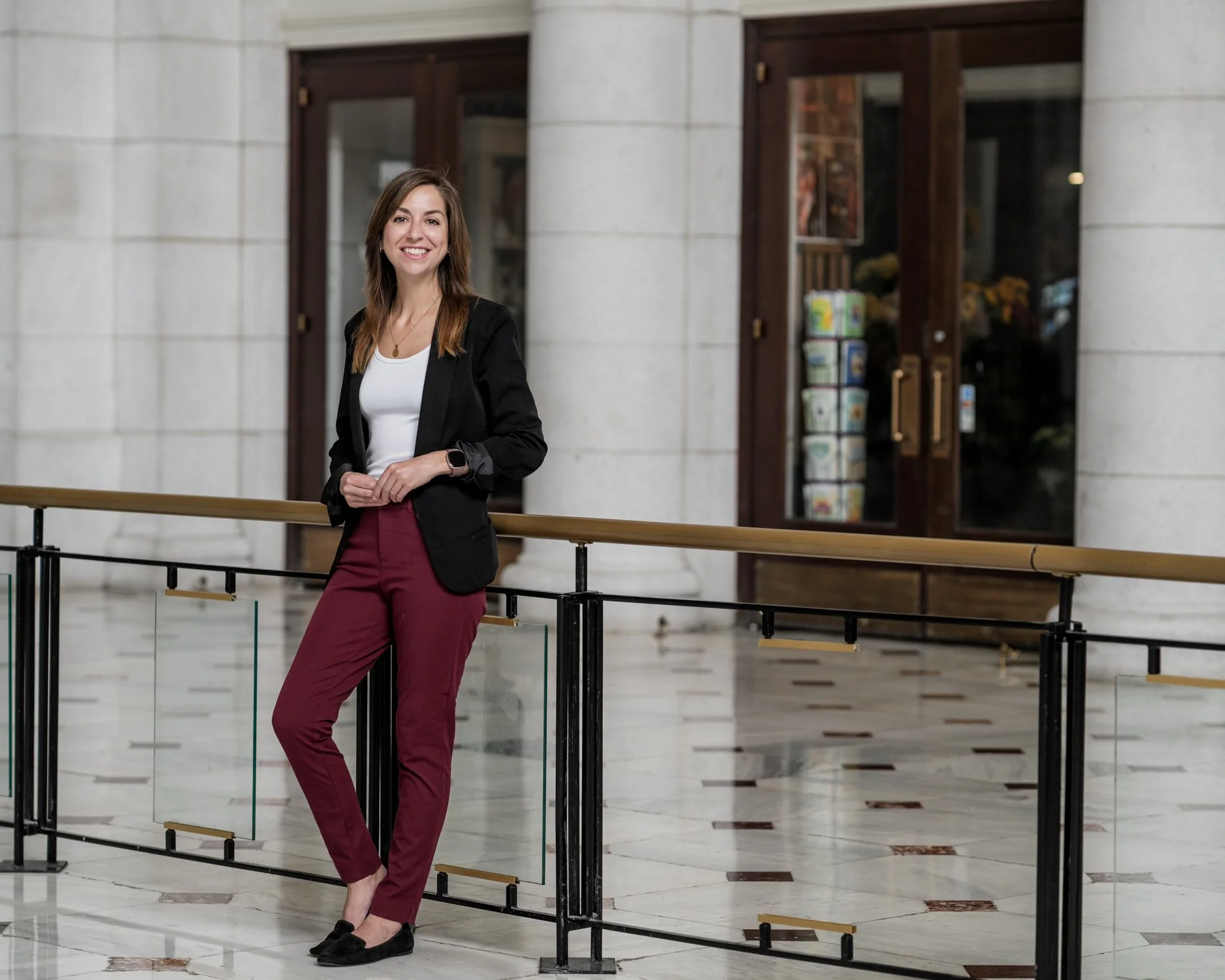 A smiling woman in business attire standing inside a building, leaning against a railing in front of a large front door.