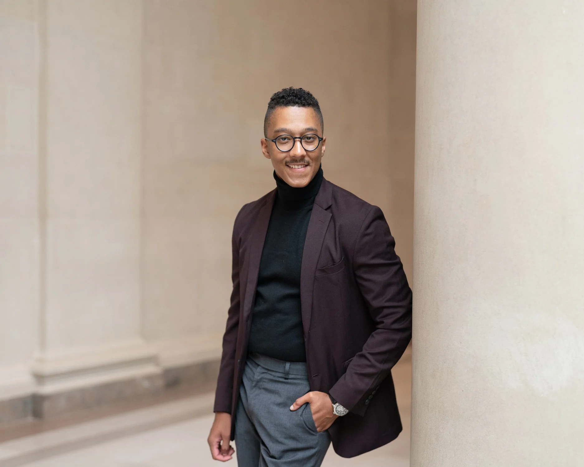 A man in a black turtleneck, dark blazer, and glasses standing against a beige pillar, smiling and looking at the camera.