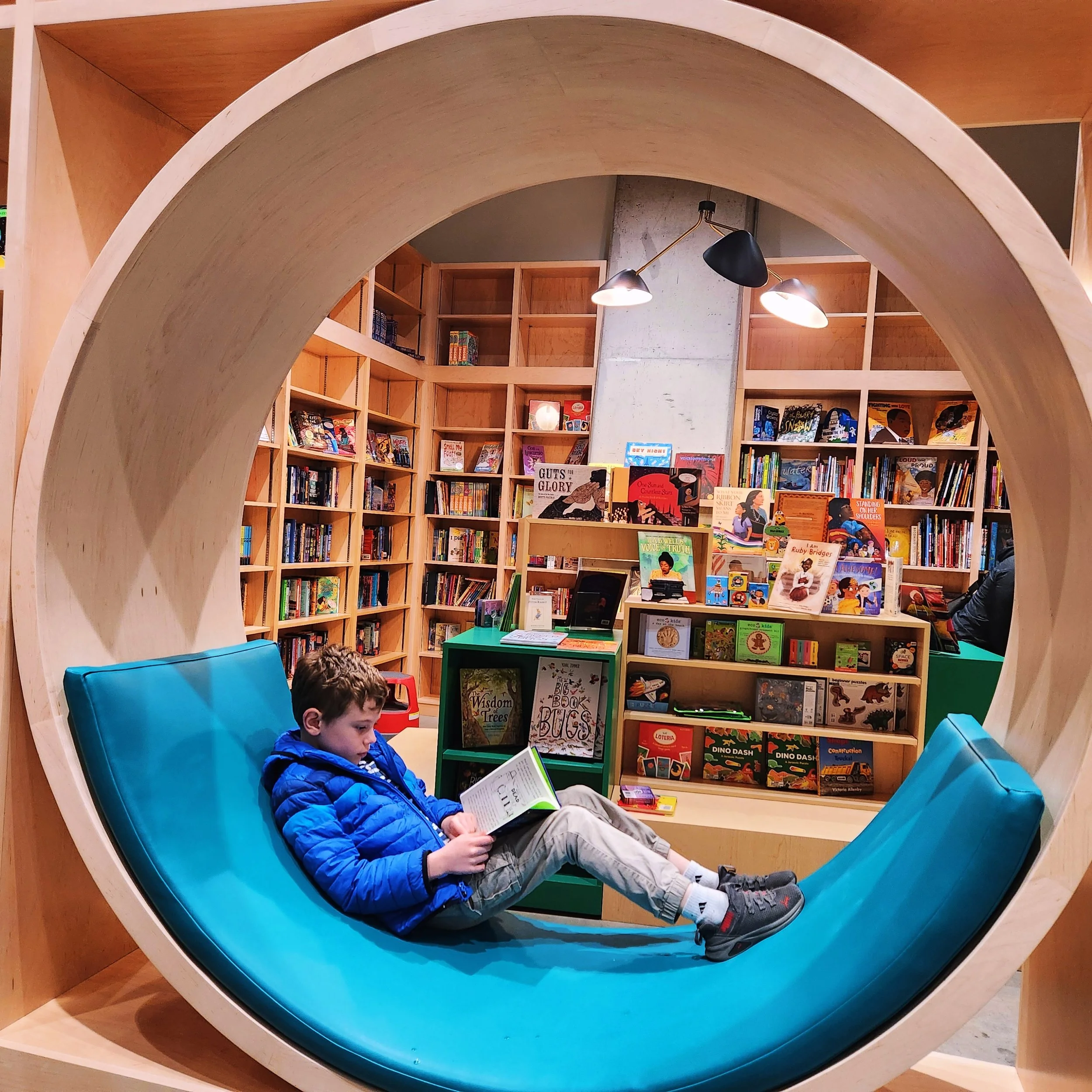 A young boy reading a book while lying on a curved bright blue cushioned seat inside a bookstore.