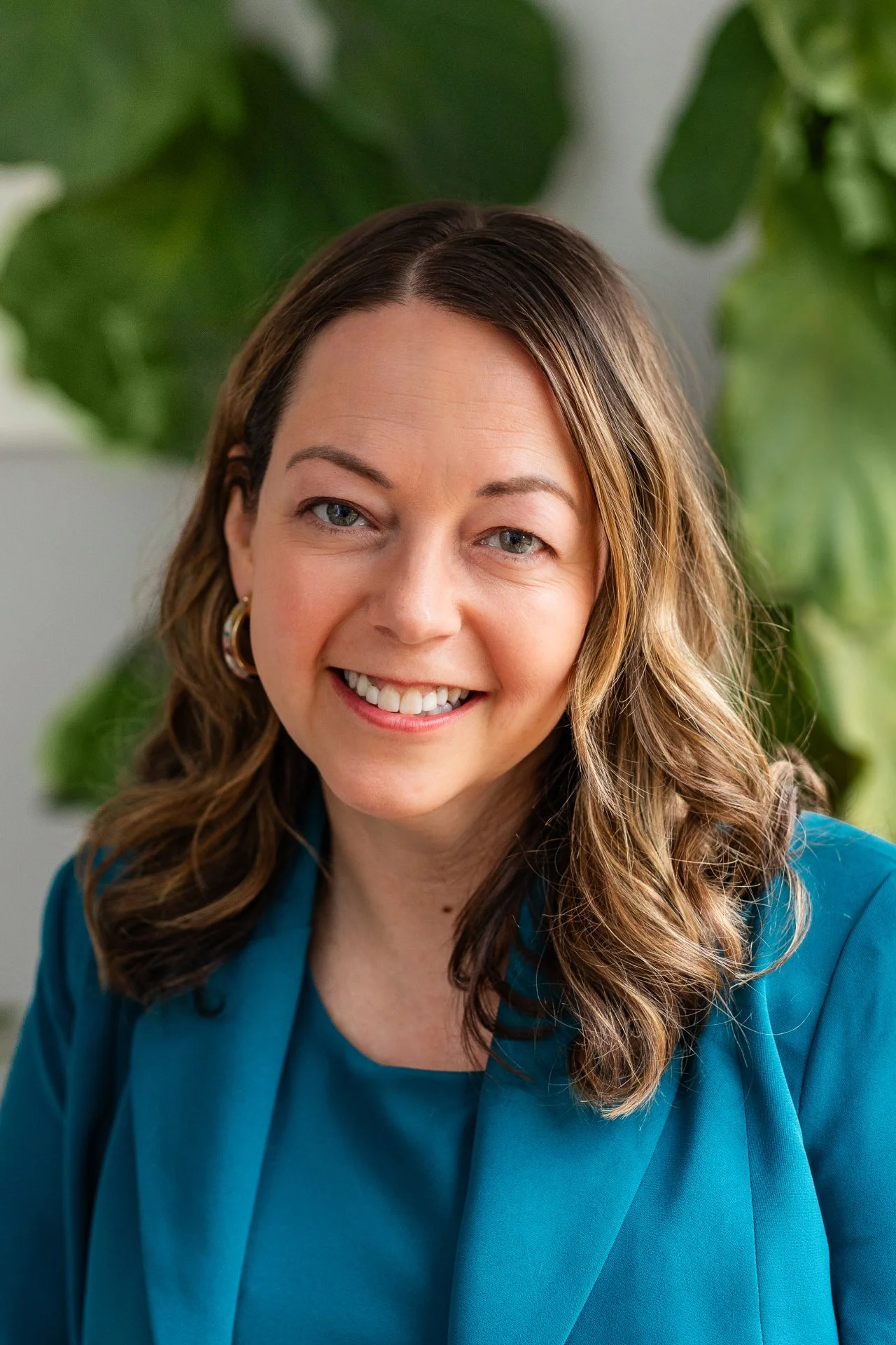 A woman with shoulder-length brown hair, blue eyes, and a bright smile wearing a teal blazer and top, standing in front of green leaves.
