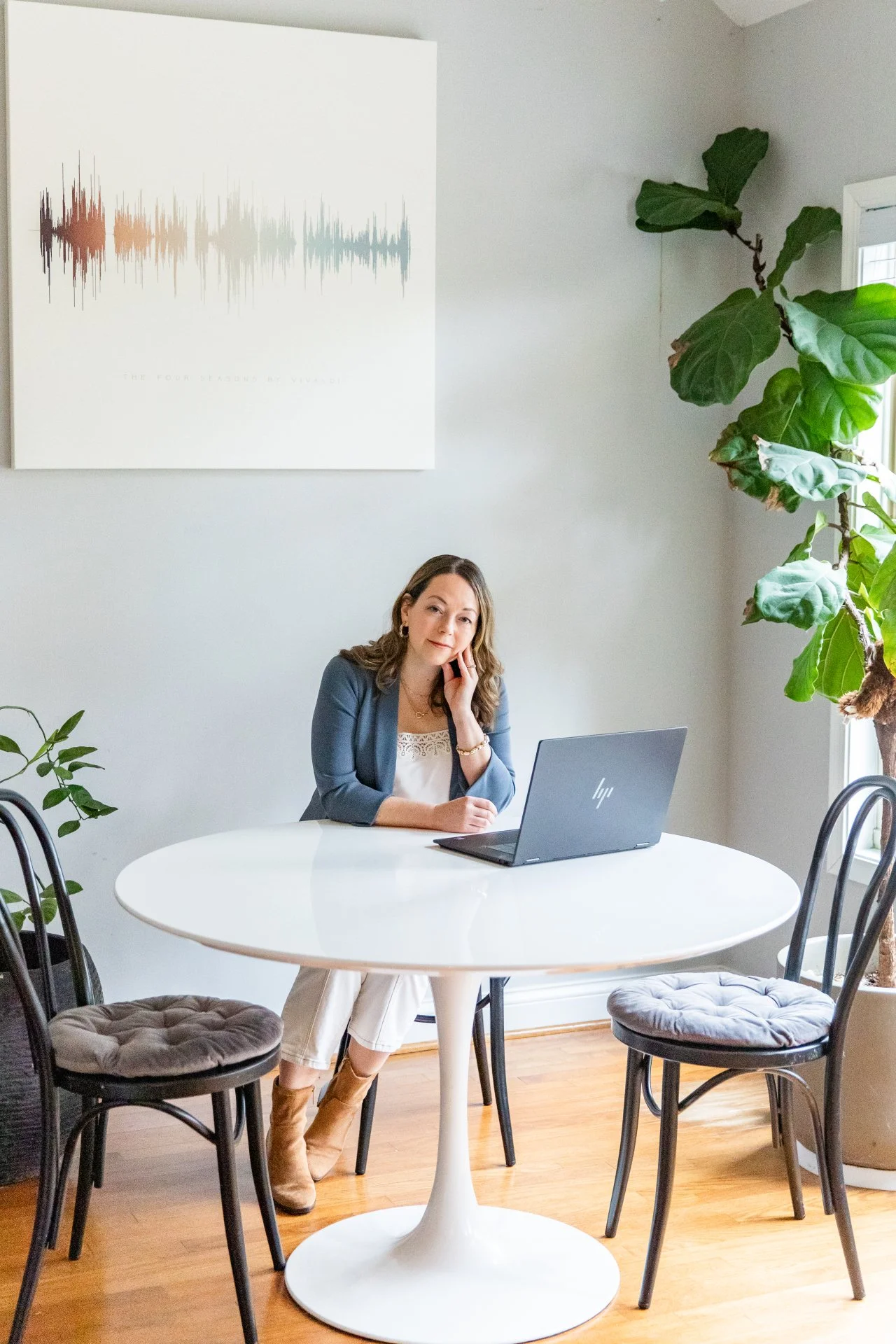 A woman sitting at a round white table with a laptop, inside a room with light-colored walls, large green plants, and a wall art piece with soundwave design.