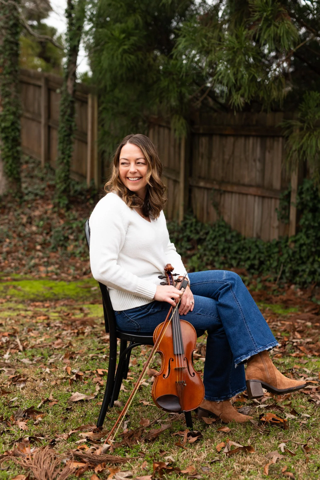A woman sitting outdoors on a black chair holding a violin and bow, smiling and looking to the side, with a wooden fence and green trees in the background.