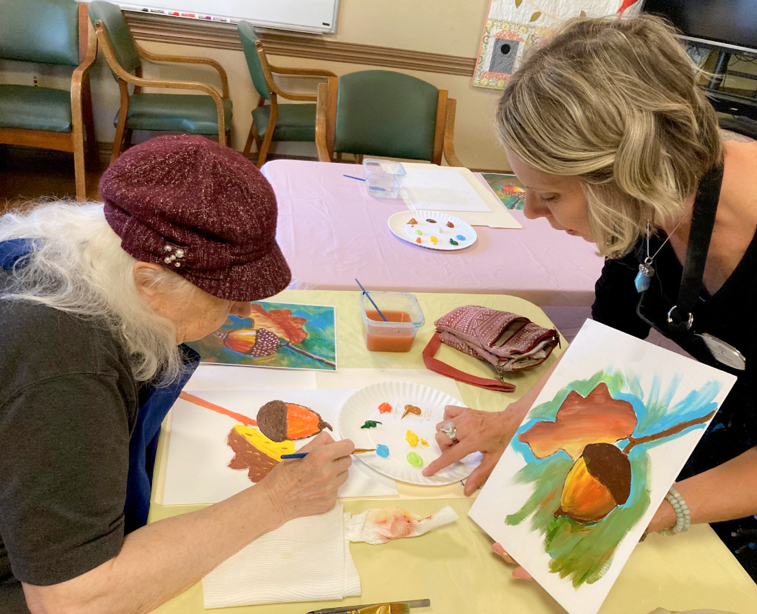 An older woman and a younger woman are painting autumn-themed pictures of trees with colorful leaves on paper at a table. The woman on the right is holding a painting, and the woman on the left is working on her own. There are paint supplies, a plate with paint, and a small container of water on the table.