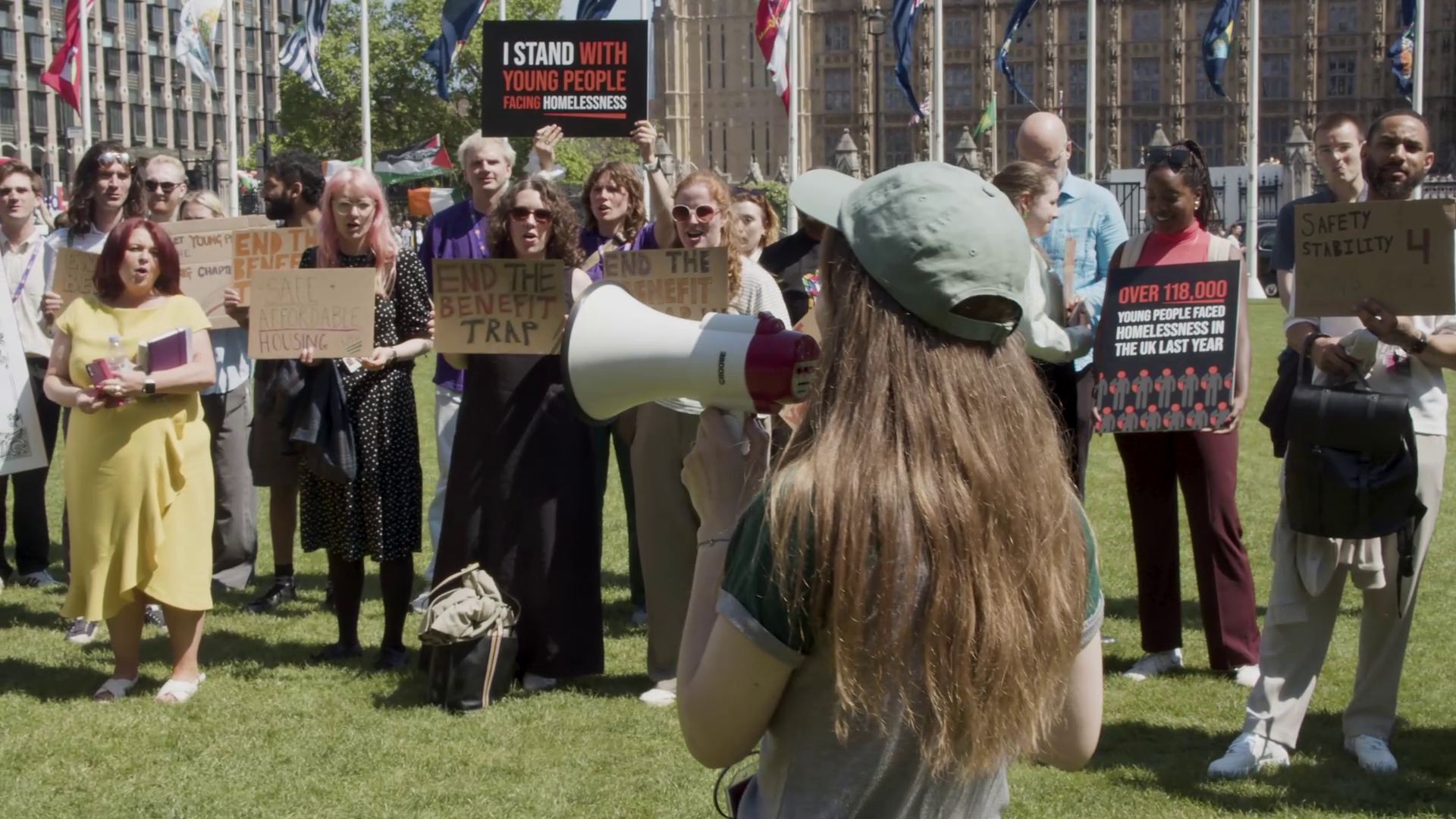 A group of campaigners outside parliament holding signs and banners. In the centre of the image is someone with a megaphone