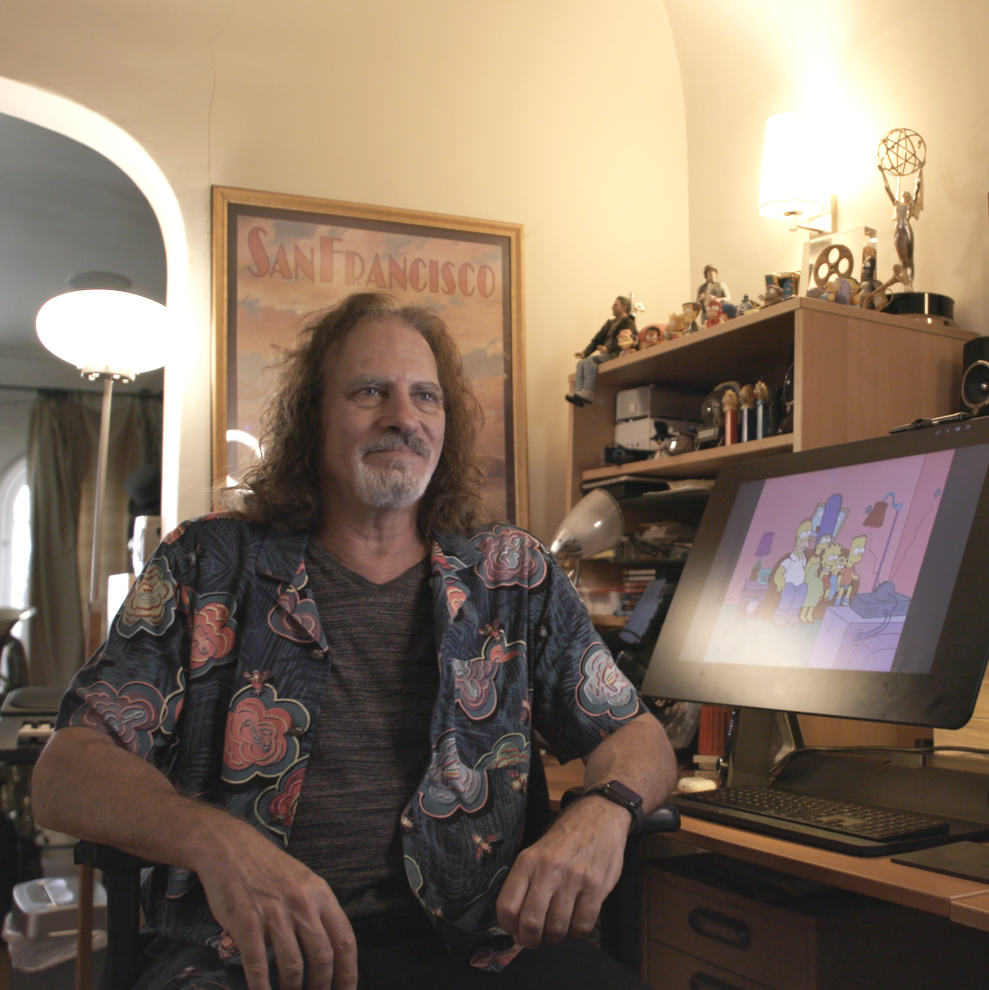 A man with long curly hair and a beard sitting at a desk with a computer monitor displaying The Simpsons. The room has a bookshelf with toys and memorabilia, a framed 'San Francisco' poster, and lamps providing warm lighting.