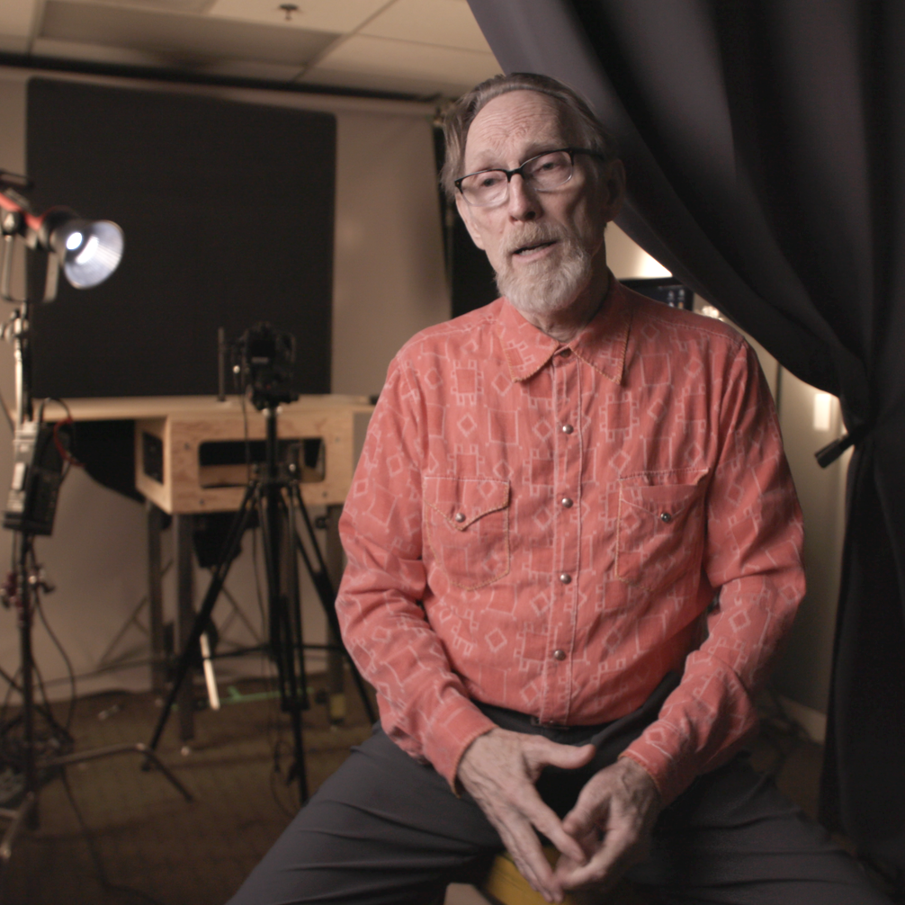 A man with glasses and a beard sitting in a recording studio with cameras and lights around him, speaking or being filmed.