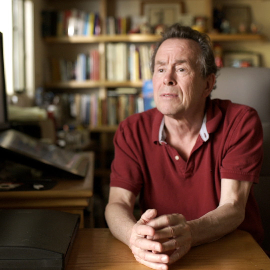 A man with gray hair and a serious expression, sitting at a desk with hands clasped in front of him, in front of a book-filled bookshelf.