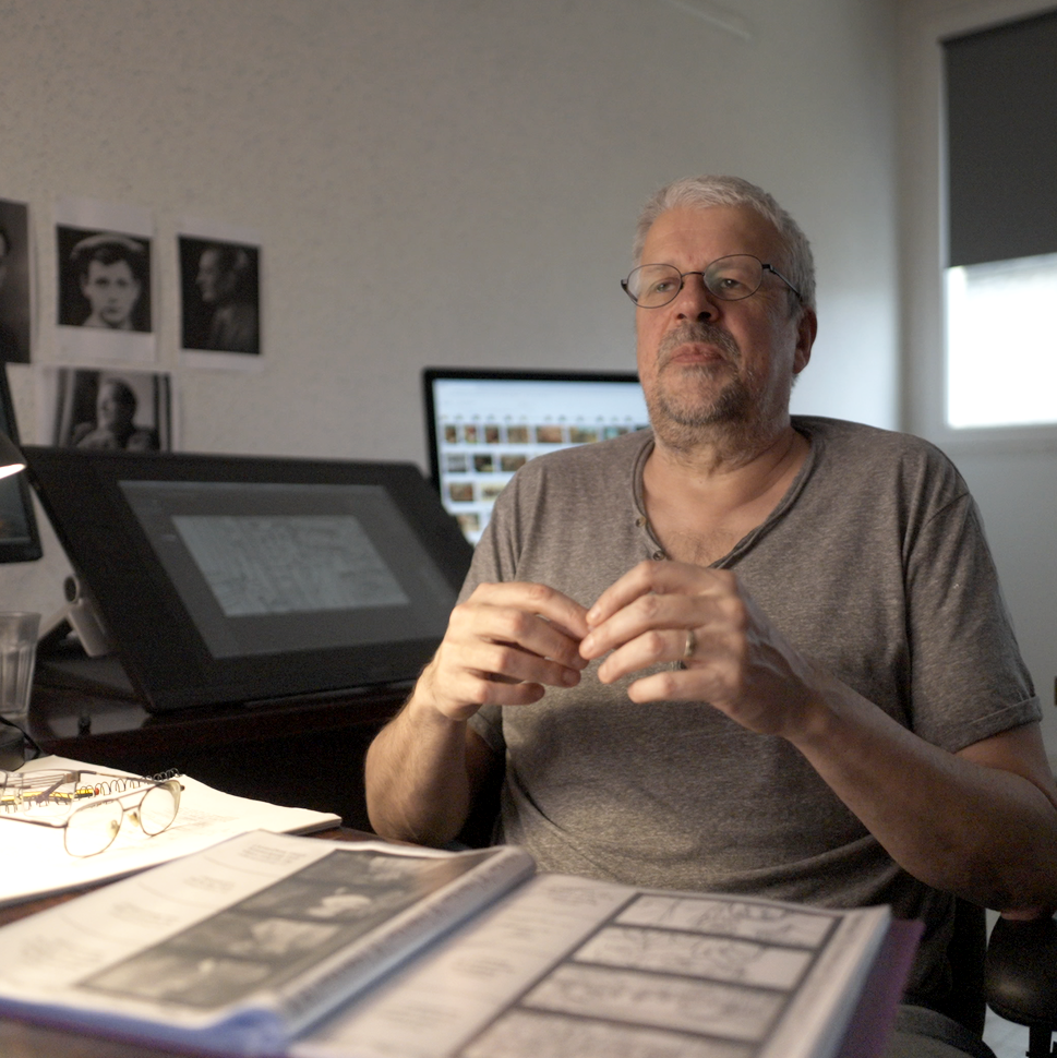 A man with glasses and a gray beard sitting at a desk in a home office, with photo prints on the wall, a computer monitor, a drawing tablet, and a book with open comic strips.