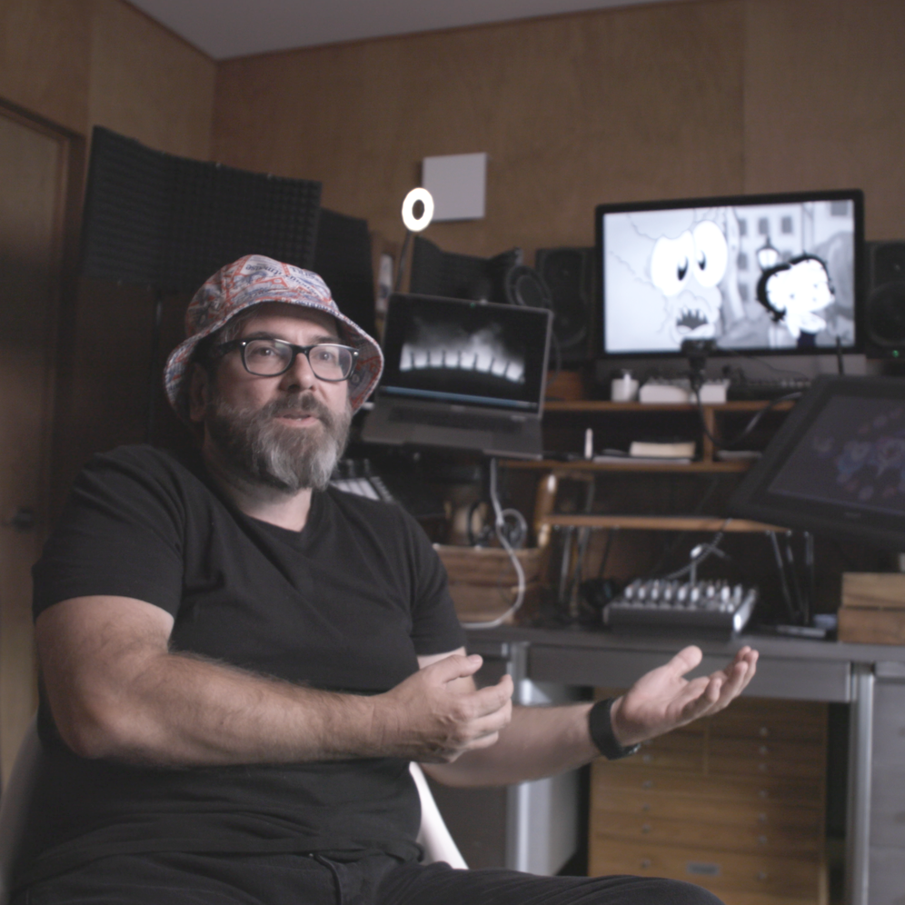A man with glasses, a beard, and a patterned bucket hat sitting in a room with multiple monitors, some displaying animations, and audio equipment.