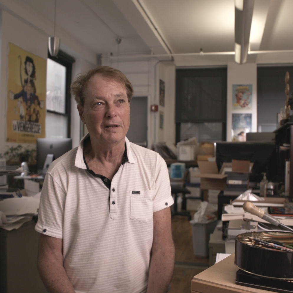A man with light brown hair standing in an office or studio space, looking thoughtful, with posters and various office equipment around him.
