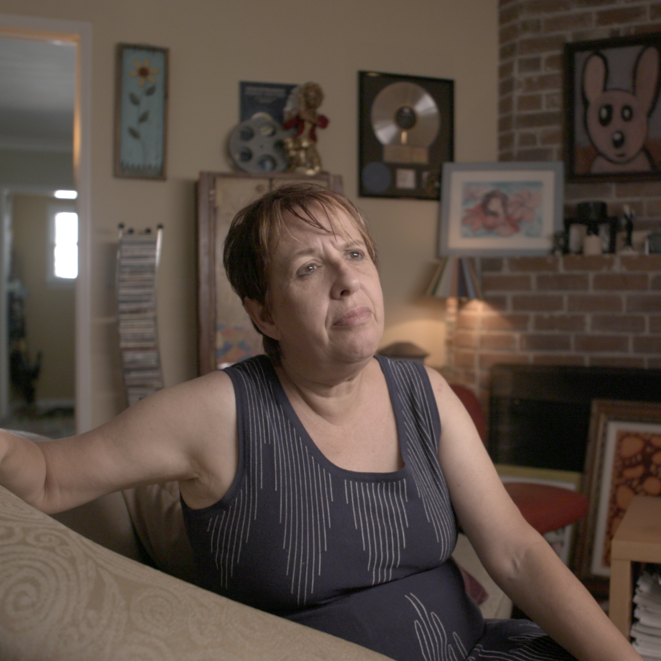 A woman sitting on a sofa in a living room, with framed artwork and awards on the wall behind her.