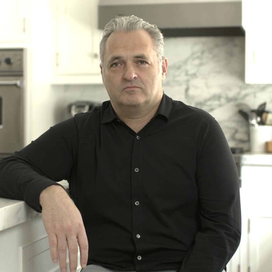 A man with gray hair wearing a black shirt, sitting in a kitchen with white cabinets and a marble backsplash.