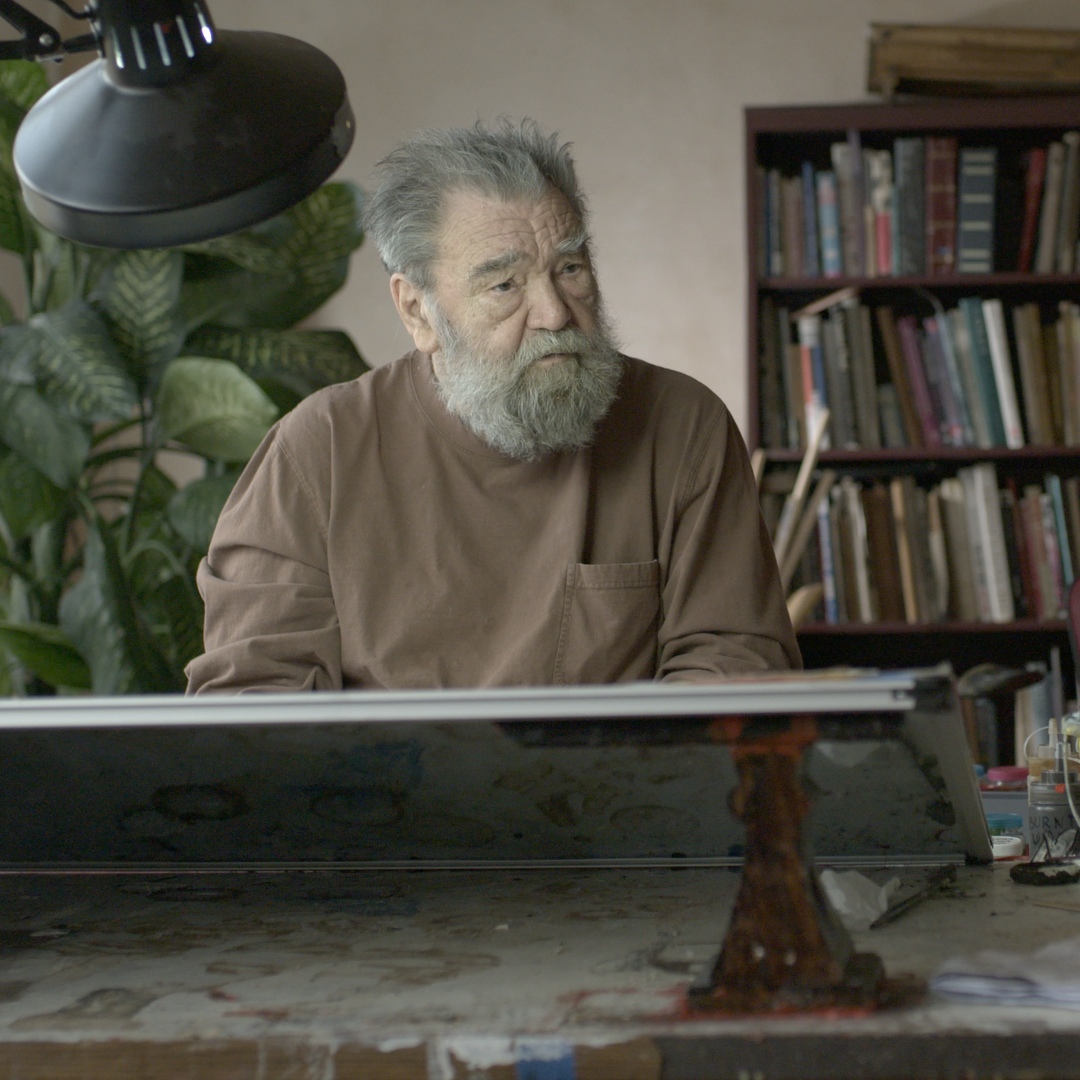 A man with gray hair and a beard sitting at a desk, looking thoughtful, with a large plant and a bookshelf in the background.
