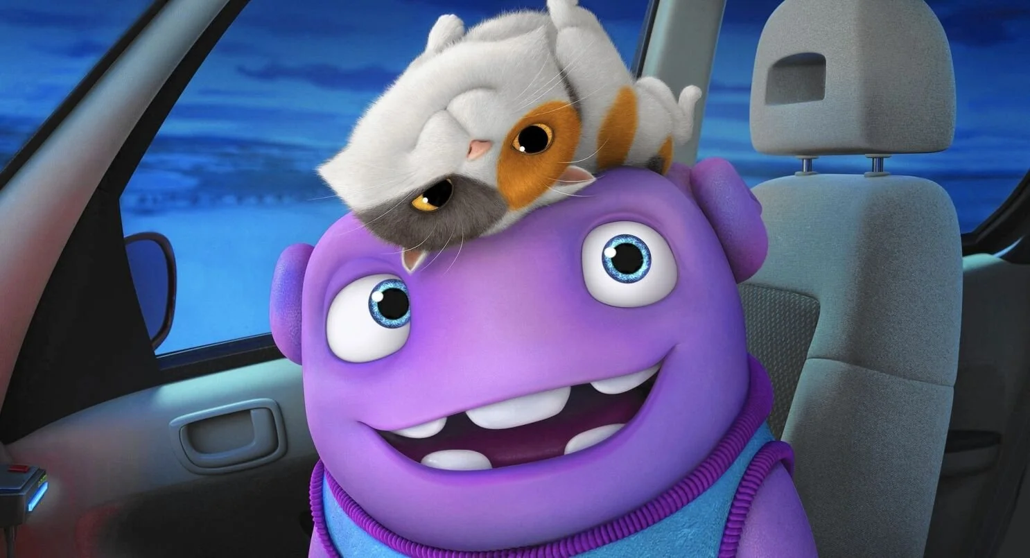 A cartoon from Home of an alien with big blue eyes and a wide smile sitting in a car seat, with a cat lying on its forehead inside a vehicle, with a landscape visible outside the window.