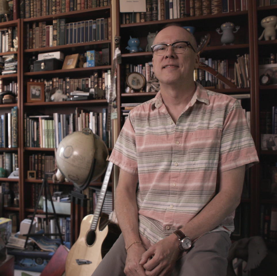 A man with glasses and a striped short-sleeve shirt sitting in front of a large bookshelf filled with books and collectibles.