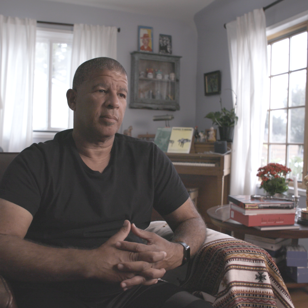 Man sitting on a couch in a cozy living room with natural light, a piano, books, and houseplants in the background.