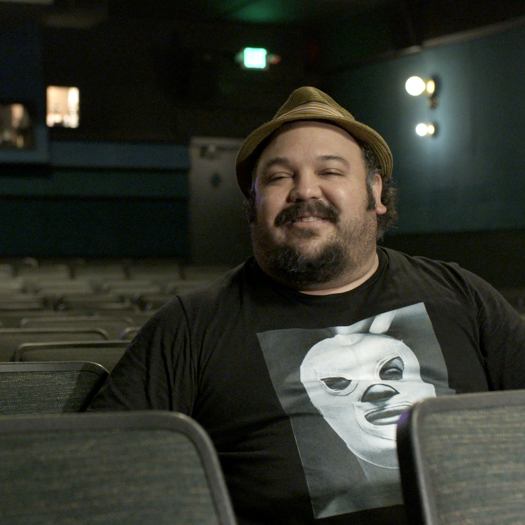 A man smiling and sitting in a theater, wearing a hat, a black t-shirt with a large graphic.