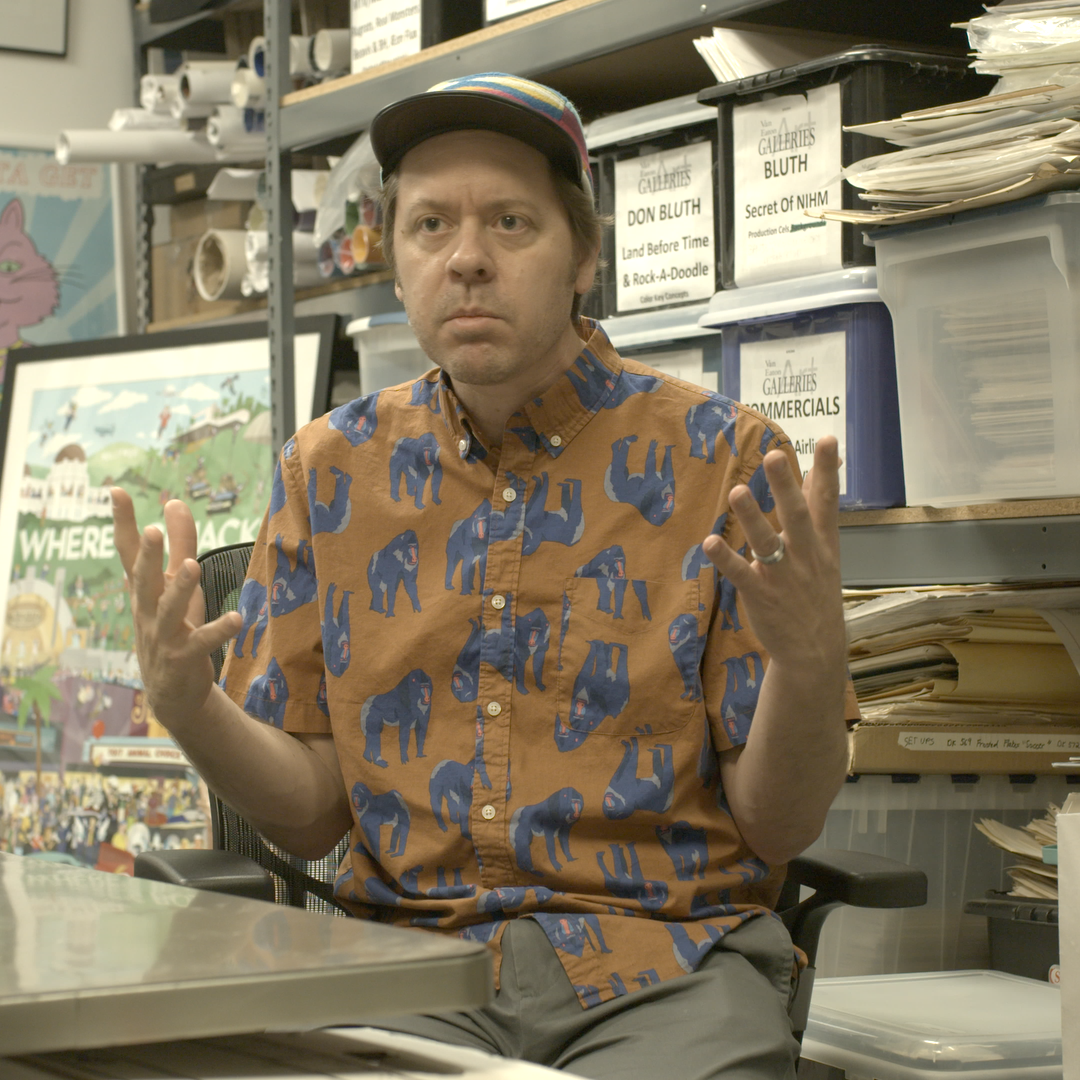 A man wearing a patterned short-sleeve shirt and a baseball cap, sitting at a desk with shelves of files and boxes behind him, gesturing with his hands.