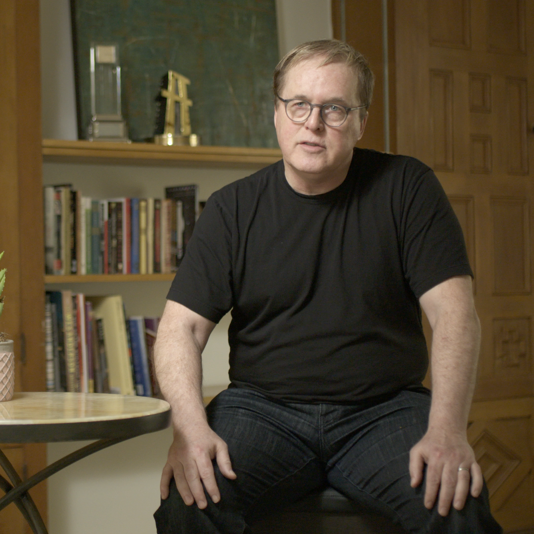 A man with glasses and a black t-shirt sitting on a stool in a room with wooden furniture and a bookshelf in the background.