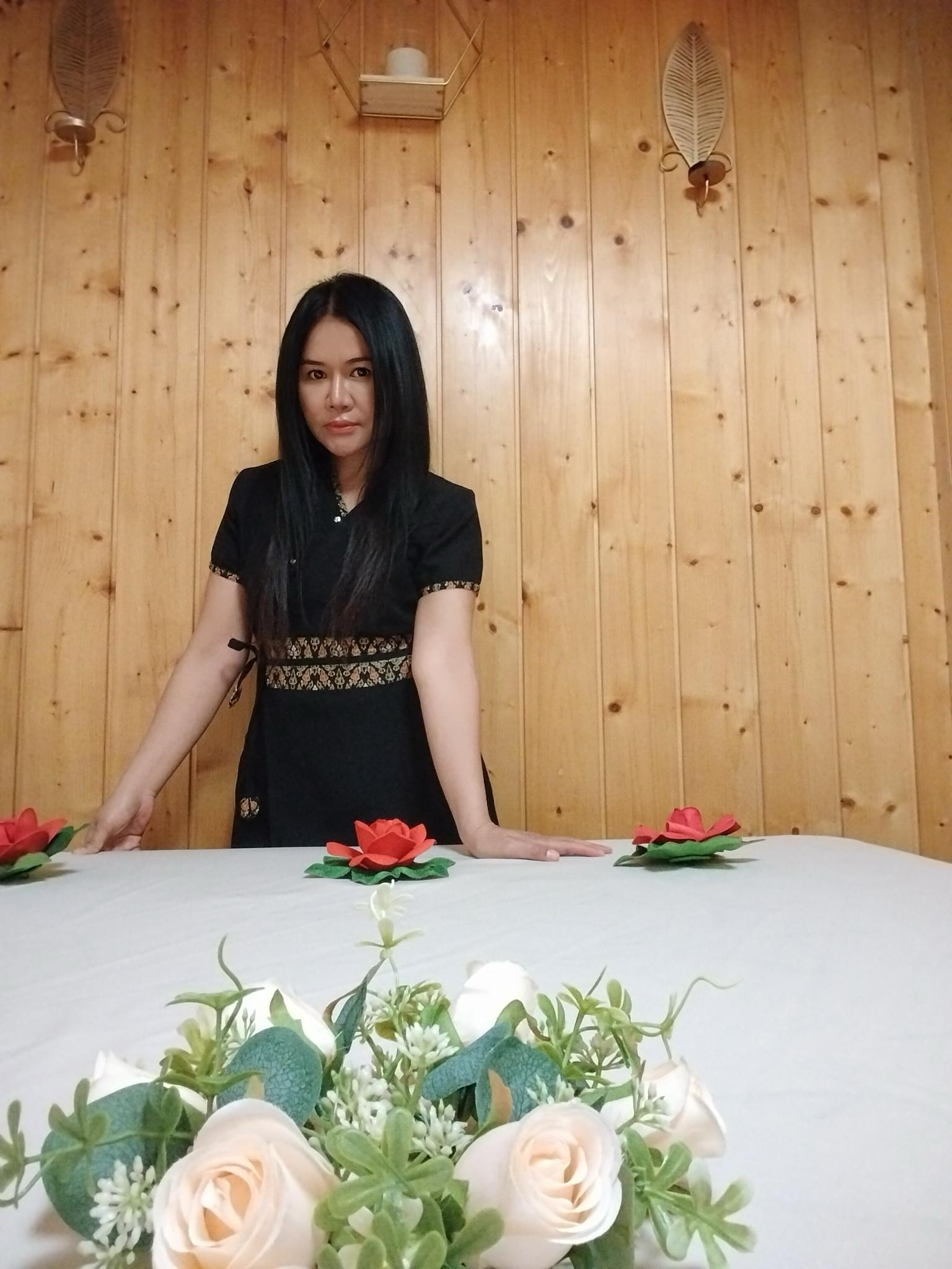 Eine Frau mit schwarzen Haaren lehnt an einem weißen Tisch, der mit weißen und grünlichen Blumen dekoriert ist, vor einer Holzvertäfelung. Auf dem Tisch sind auch rote Papierblumen. An der Wand sind zwei metallene Wandlampen und eine kleine weiße Ablage mit einem Gegenstand drauf.