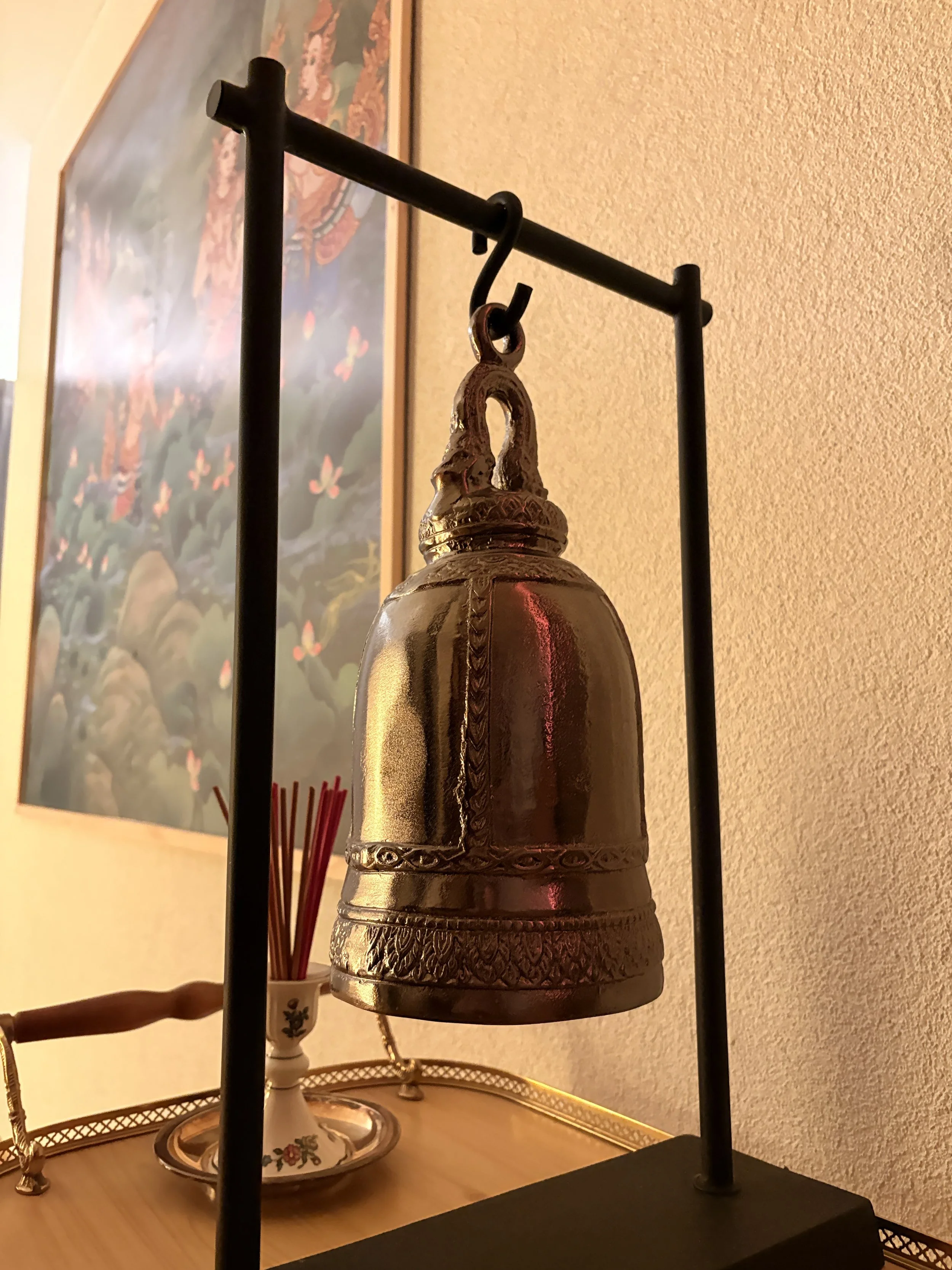 Ein goldener, verzierter Glocke an einem schwarzen Metallgestell. Im Hintergrund eine Vase mit roten Stäbchen und ein Wandbild mit Blumen.