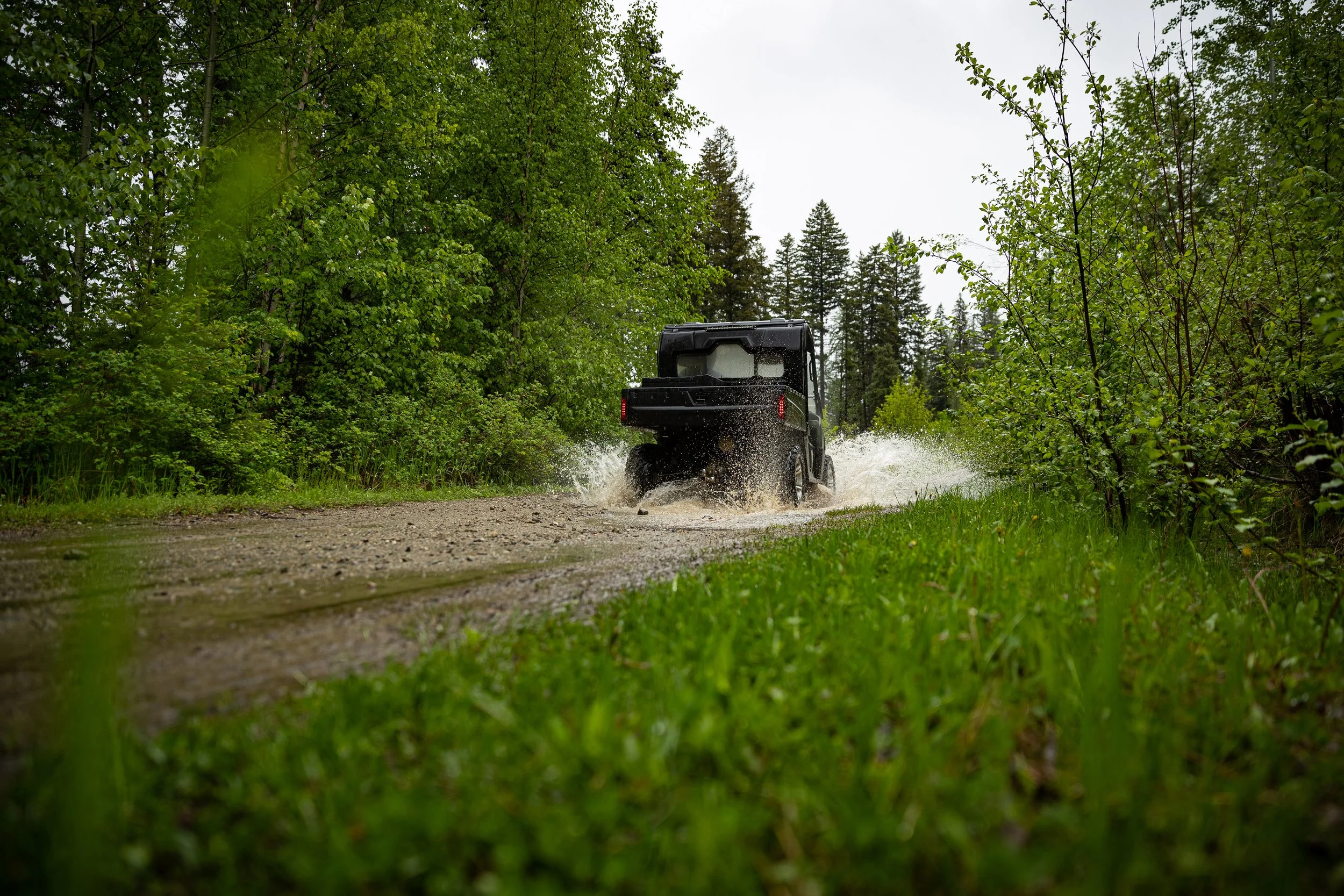 A black off-road vehicle driving through a muddy trail in a forest with dense green foliage and trees on either side.