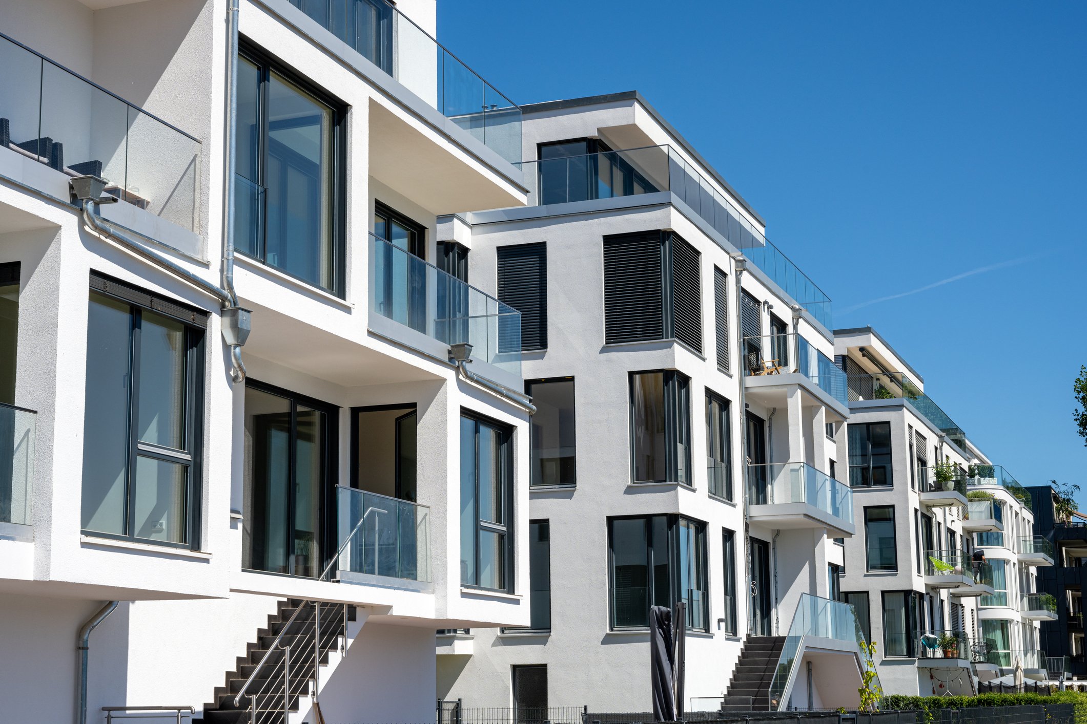 Modern white apartment building with multiple floors, large glass windows and balconies, black shutters, and stairs outside, under a clear blue sky.