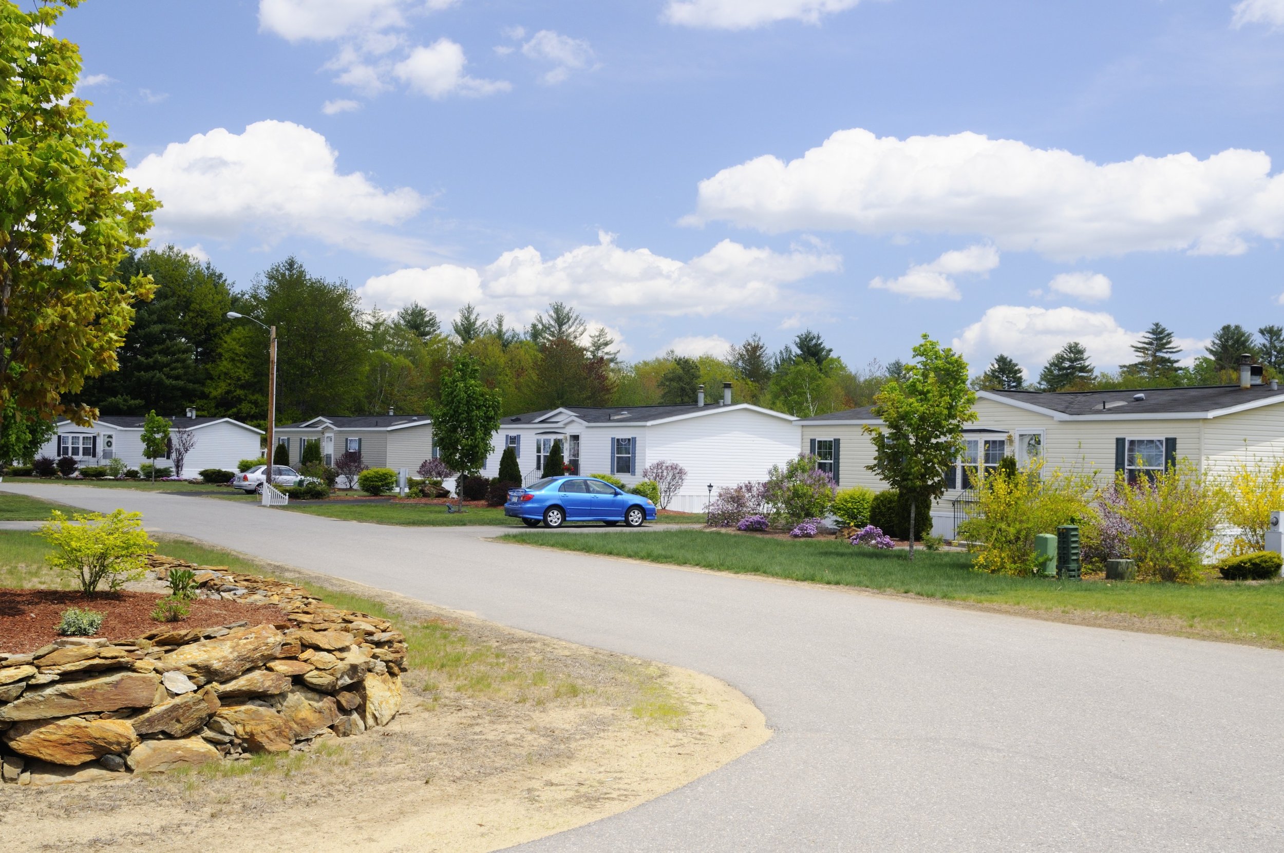 A residential neighborhood with single-story houses, a well-maintained lawn, colorful bushes, and trees. The sky is partly cloudy with fluffy white clouds and a blue sky.