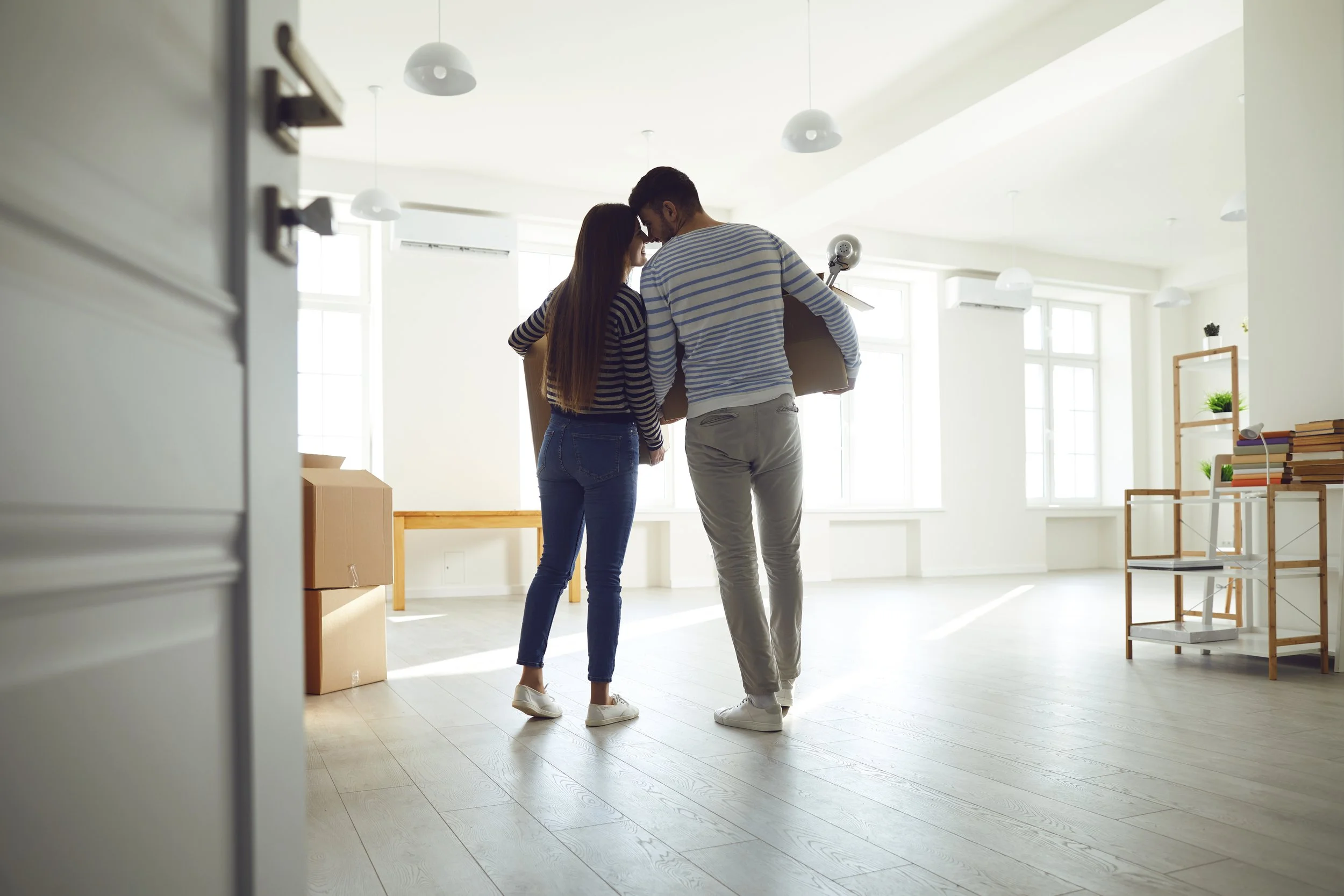 A young couple with boxes in a bright, spacious room with large windows, light wood flooring, and minimal decor.