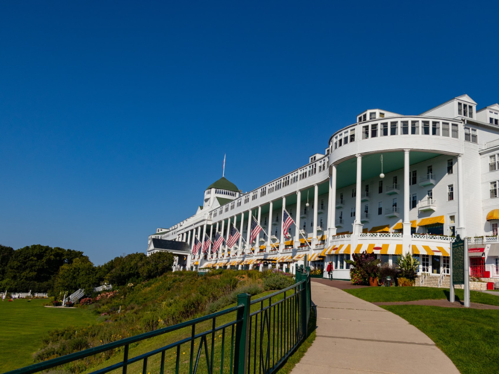 Large white historic hotel building with a curved facade, multiple stories, yellow and white awnings, and multiple American flags, located near a well-kept lawn with a walkway.