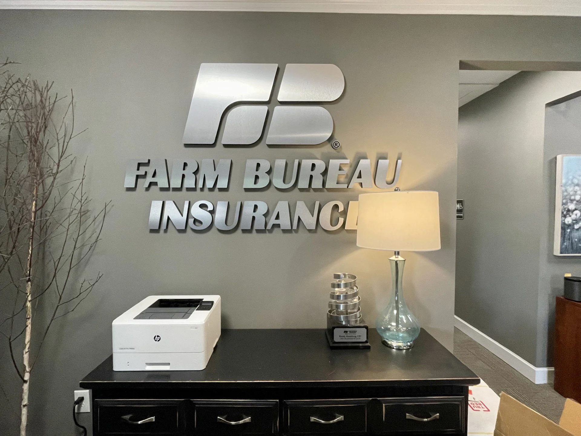 Interior of an office with a wall sign reading 'FARM BUREAU INSURANCE' with a logo, a table with a white printer, a decorative glass vase, a lamp, and surrounding furniture.