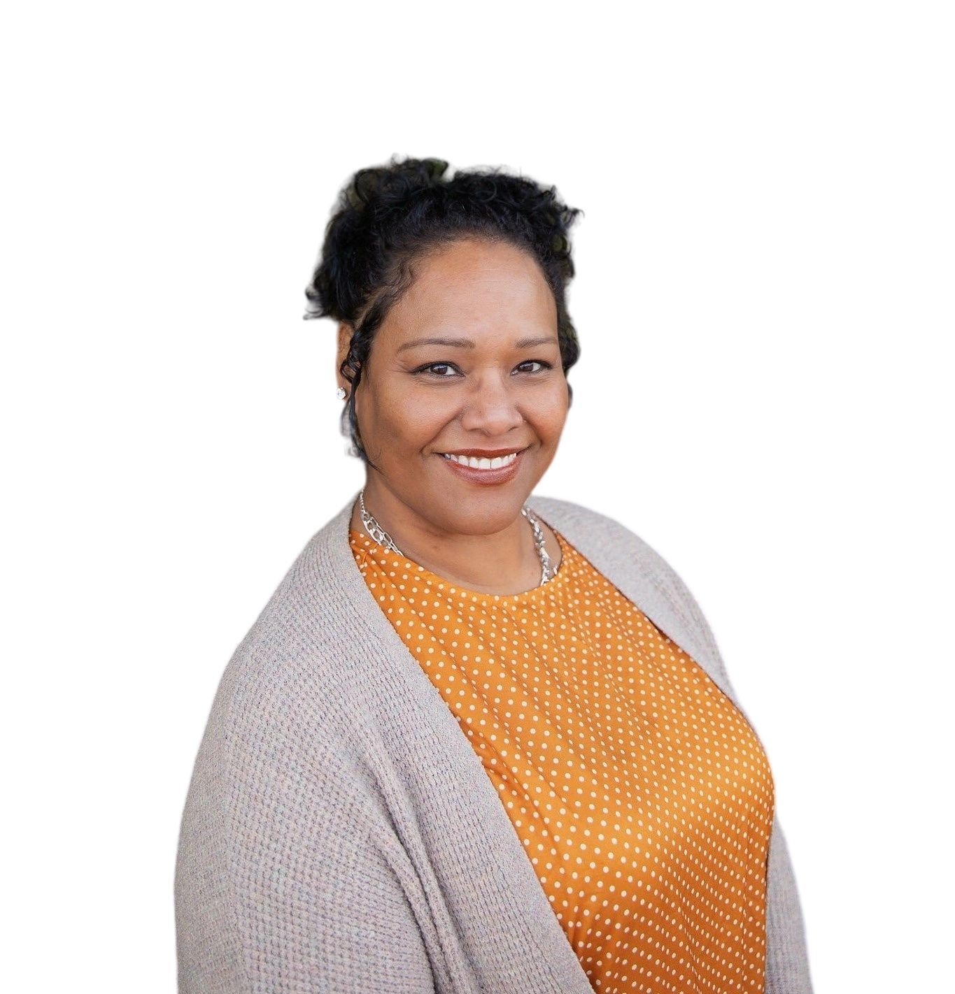Portrait of a smiling Black woman wearing a beige cardigan and orange polka dot blouse, with short curly black hair and jewelry, against a white background.