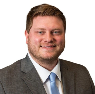 A man with short brown hair, a beard, wearing a gray suit, white shirt, and light blue tie, smiling against a white background.